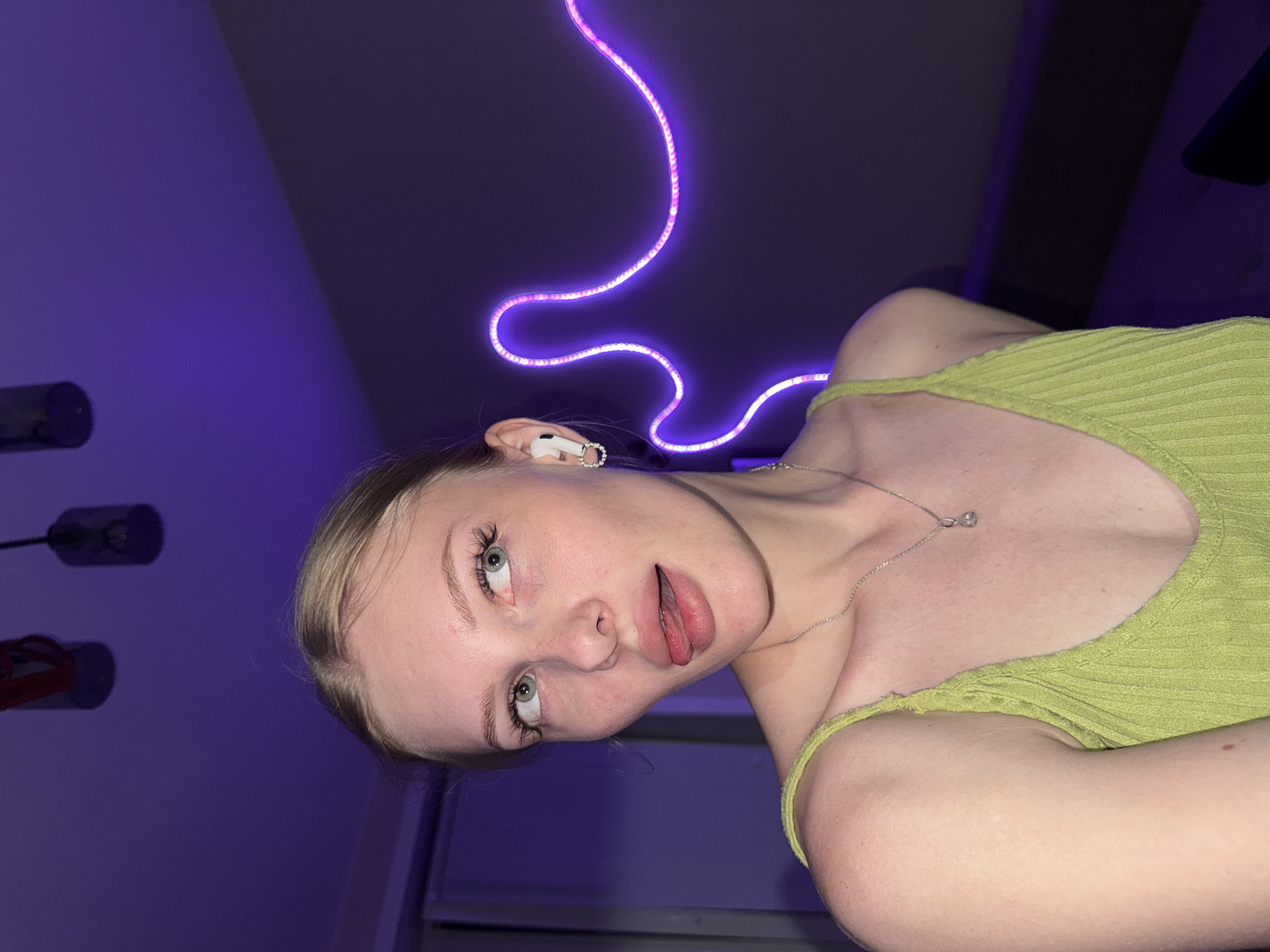 A young Caucasian woman with blonde hair pulled back and green eyes is looking to her left with her tongue sticking out. She is wearing a green ribbed tank top and a silver necklace with a diamond pendant. She has white wireless earbuds in her ears. Behind her, a purple neon light strip forms a wavy pattern on a dark gray wall. Two pendant lights hang from the ceiling above her. The scene appears to be indoors, possibly in a bedroom, with a dresser or closet visible on the left side of the frame. The image quality is high, with good lighting and focus. The location is not specified to be in the city of Perm, Russia.