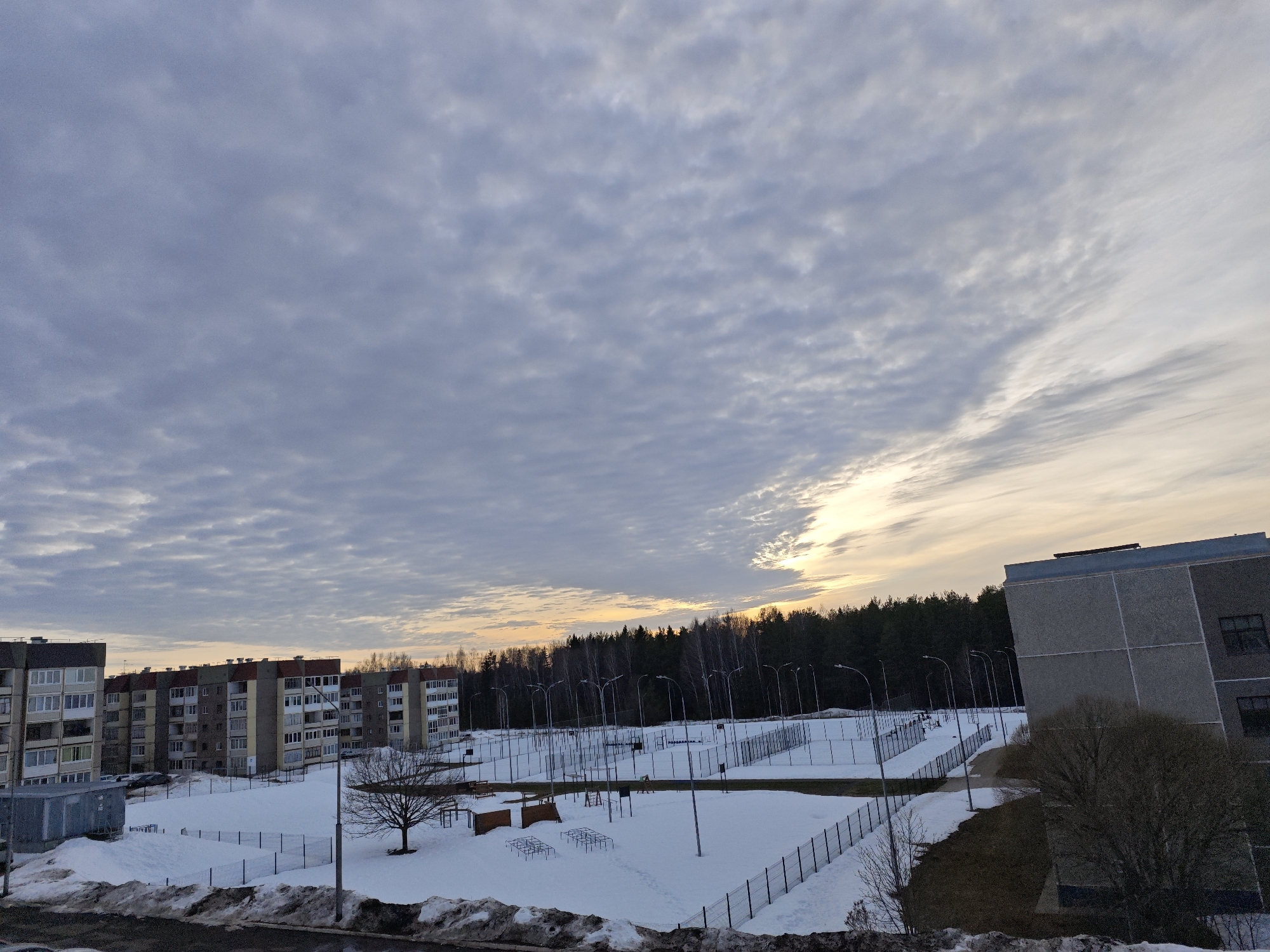 The scene depicts an outdoor urban environment in Markovskii, Russia, under a heavily clouded sky at twilight, exhibiting characteristics of late winter or early spring. The sky is dominated by a dense layer of altocumulus clouds, transitioning to a bright yellow and orange luminescence on the right horizon. The ground is extensively covered in snow, with patches of exposed brown earth and dark, melted snow accumulation visible in the immediate foreground and along pathways.
To the left, a series of multi-story, residential-style apartment buildings with light-colored facades, multiple windows, and balconies are visible. Several vehicles are parked in the snow-covered areas adjacent to these structures. A small, gray metallic utility box is positioned in the lower left corner.
The central and mid-ground is occupied by a large, snow-covered recreational or sports complex, enclosed by dark metal fencing. This area features multiple sports courts or fields, delineated by snow-covered markings and structures. Numerous tall, dark streetlights with single luminaires are systematically arranged throughout the complex. Several pieces of outdoor fitness or playground equipment, including metallic frames resembling monkey bars, are partially visible through the snow. A solitary, bare deciduous tree stands within this complex.
In the background, a dense line of evergreen forest forms the horizon. To the far right, a large, multi-story building constructed with gray concrete panels is partially visible, featuring a flat roof and dark-framed windows. Several dormant deciduous trees and shrubs are located in front of this building and along an elevated earth bank. No individuals are present within the captured scene.
