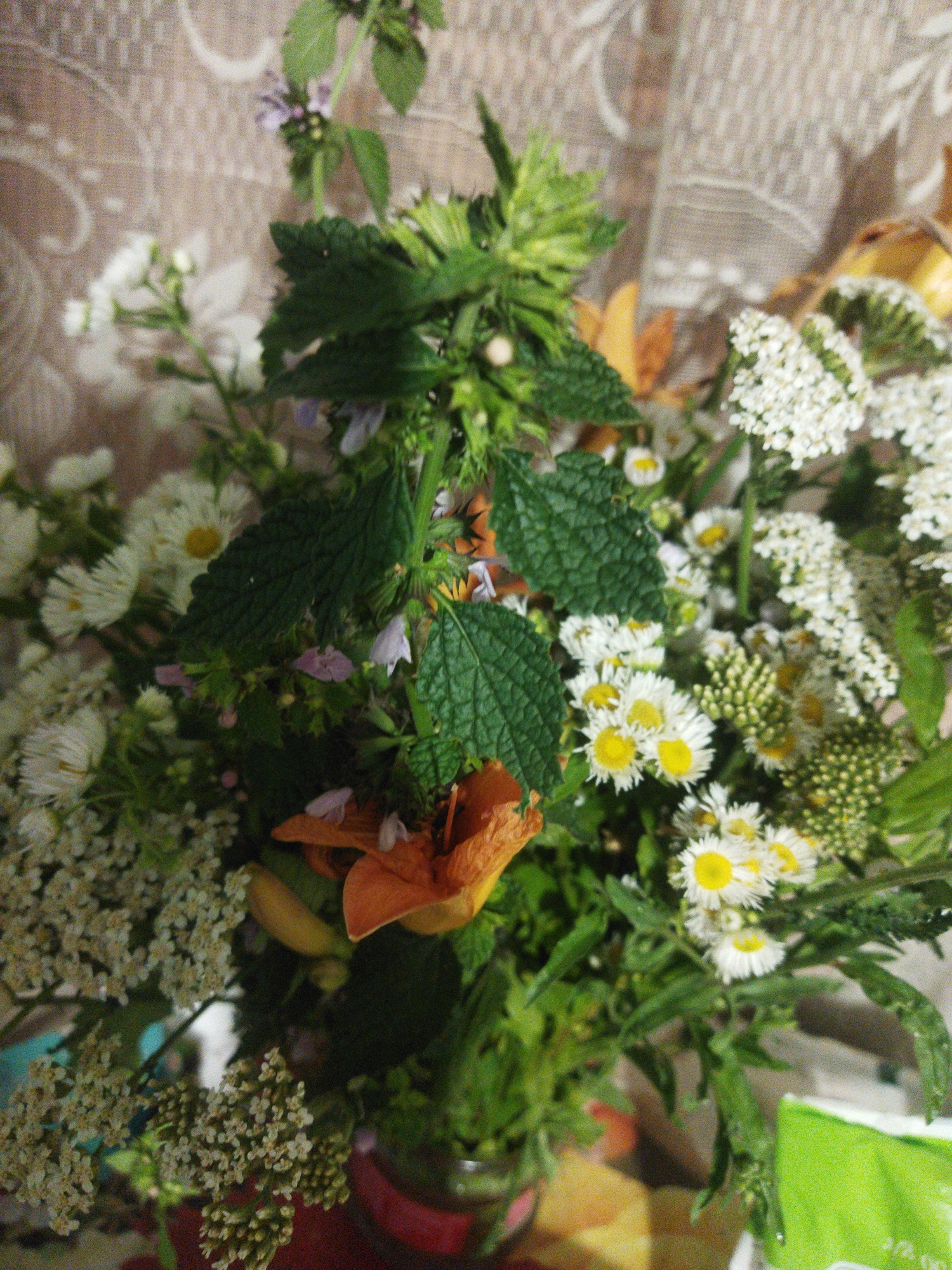 A bouquet of wild flowers is displayed in a vase against a patterned backdrop. The bouquet contains a variety of flowers including white daisies, small white clusters, and a few orange lilies. Green leaves and stems are interspersed throughout the arrangement. The background is a light brown fabric with an embossed pattern. The image is a close-up shot, with the flowers filling the majority of the frame. The exact location in the city of Sosnivka, Ukraine cannot be determined from the visual information.