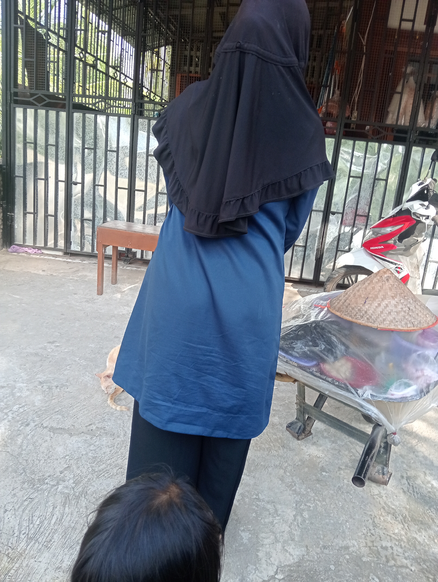 In Campago, Indonesia, a person wearing a black hijab and a blue tunic stands on a concrete ground outside a building with a metal fence. A child's head is visible in the foreground. Behind the person is a wooden table. To the right, a motorcycle and a cart are visible. A light-colored cat is lying on the ground to the left of the person. The environment appears to be an outdoor area, possibly a yard or a street.