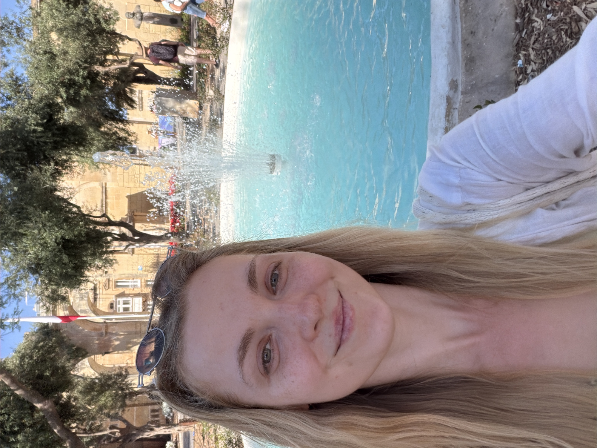 This media file is a portrait-oriented photograph featuring a young woman taking a selfie in front of a fountain. The woman has long blonde hair and is smiling, with sunglasses resting on her head. She is wearing a white top, and her expression suggests a happy and relaxed mood.
The background depicts an outdoor scene with a bright blue fountain in the foreground, actively spraying water. Behind the fountain, there are trees with lush green foliage and a historic building constructed of light-colored stone. The architecture of the building, with its arched entrances and windows, hints at a Mediterranean or European location. The presence of a flagpole with a red and white flag, and the characteristic stone buildings, strongly suggest the location is Valletta, Malta. The sunlight and the clear blue water indicate a bright, sunny day, likely during daytime. In the distance, a few other people can be seen, one of whom is wearing a backpack. The overall setting appears to be a public square or park, likely a tourist attraction given the historical architecture and fountain. There is no visible text in the image.