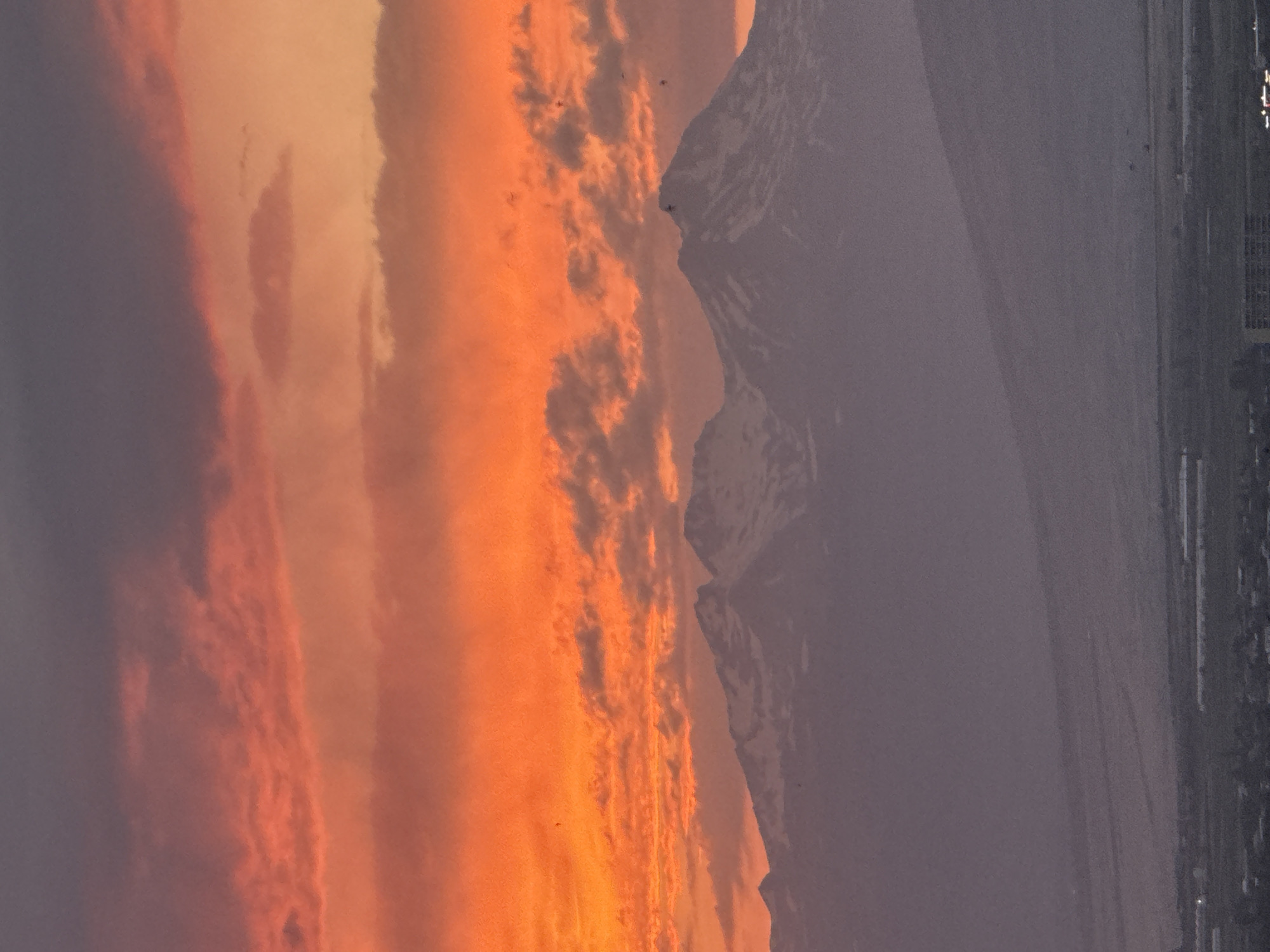The image captures a dramatic sunset over the city of Abovyan, Armenia. The sky is a vibrant display of fiery orange and pink, transitioning into deeper purples and grays towards the top. Wispy clouds are illuminated by the setting sun, creating a textured and dynamic sky.
Dominating the mid-ground are the majestic, snow-capped mountains, their peaks silhouetted against the colorful sky. The mountains appear serene and imposing, suggesting a natural landscape adjacent to the urban environment.
In the foreground, the lower part of the image shows a hazy view of Abovyan. The city lights are beginning to twinkle as dusk settles. Structures, possibly residential buildings and greenhouses, are visible, hinting at the urban development in the valley. The overall atmosphere is one of natural beauty meeting urban life, with the grandeur of the mountains and the dramatic sky providing a stunning backdrop to the city.