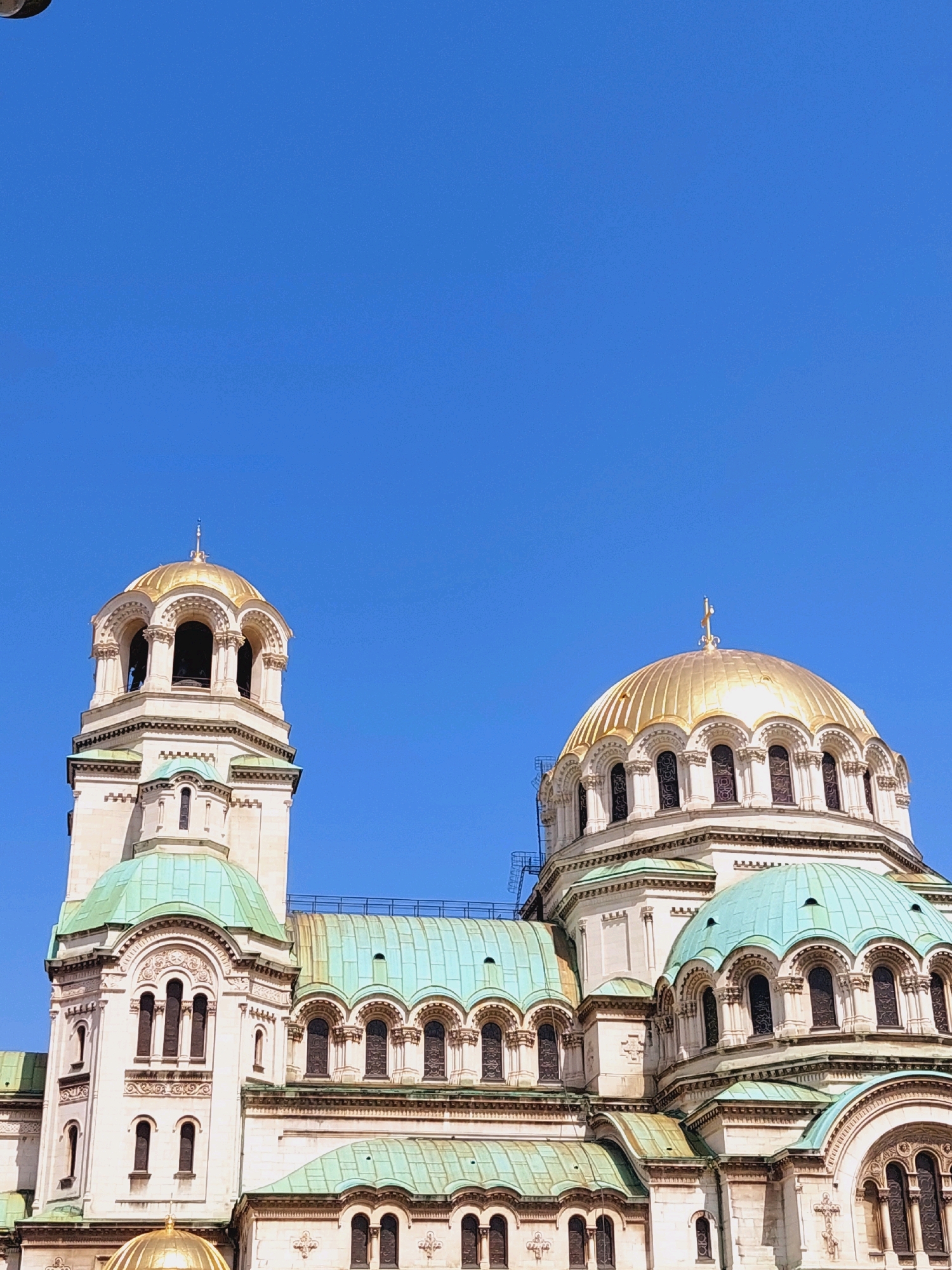 The image shows a close-up, low-angle view of the St. Alexander Nevsky Cathedral in Sofia, Bulgaria. The cathedral's dominant features are its impressive golden domes and green copper roofs, set against a clear blue sky. The architecture is characterized by intricate detailing, including arched windows and decorative carvings on the cream-colored stone facade. A prominent bell tower with a golden dome is visible on the left, while a larger, central golden dome with a cross on top is on the right. Smaller green domes and roofs are visible in the foreground and mid-ground, indicating the scale and complexity of the cathedral's structure. There are no people visible in the image. The environment is solely focused on the architectural subject, with no surrounding landscape or urban elements.