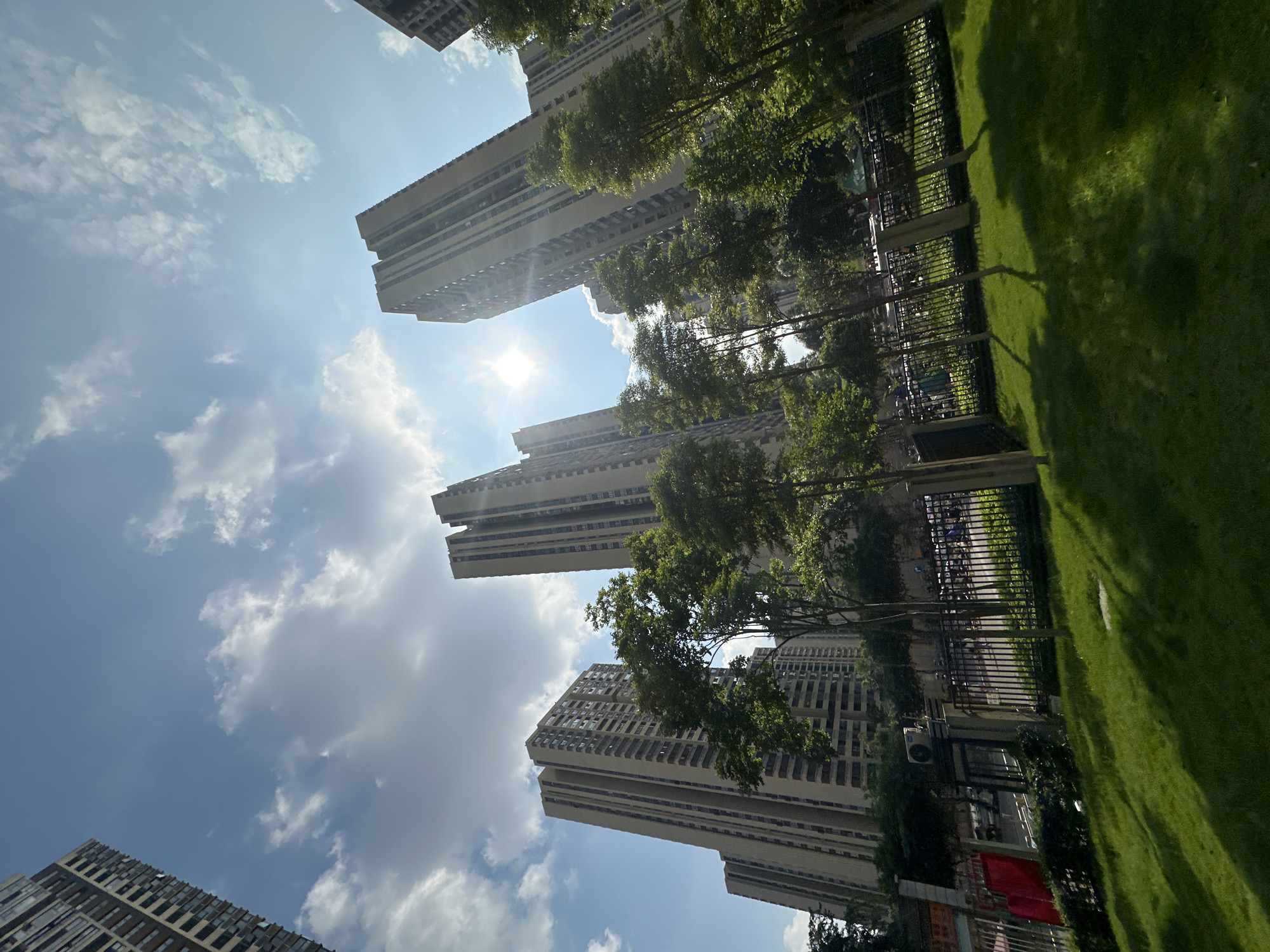 In the Chongqing High-tech Zone, China, a low-angle shot captures towering residential buildings under a partly cloudy sky. The sun is bright, creating lens flare and casting shadows on the well-maintained green lawn in the foreground. A black metal fence separates the lawn from a street where vehicles are parked. Several trees with lush green foliage are scattered throughout the scene, some in front of the buildings and others along the fence line. An outdoor air conditioning unit is visible mounted on a wall behind the fence. The buildings themselves are modern, multi-story structures with numerous windows and balconies. The overall impression is one of a contemporary urban residential area on a sunny day.