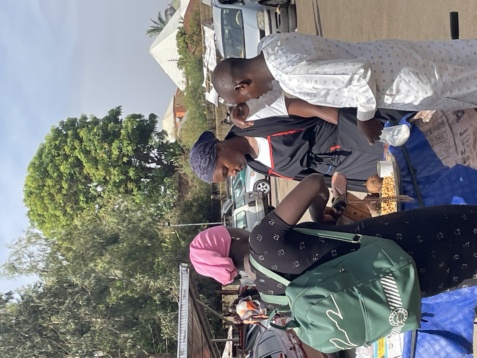In Bukuru, Nigeria, a street scene depicts three individuals at a roadside stall. A woman with a pink head covering stands beside the stall, facing the vendor. The vendor, a woman with a dark blue head covering, is attending to two male customers who are looking at items on display. One customer is wearing a white patterned shirt. The stall is covered in a blue cloth and displays food items. Cars are parked nearby, and a large tree dominates the background. The weather appears to be sunny.