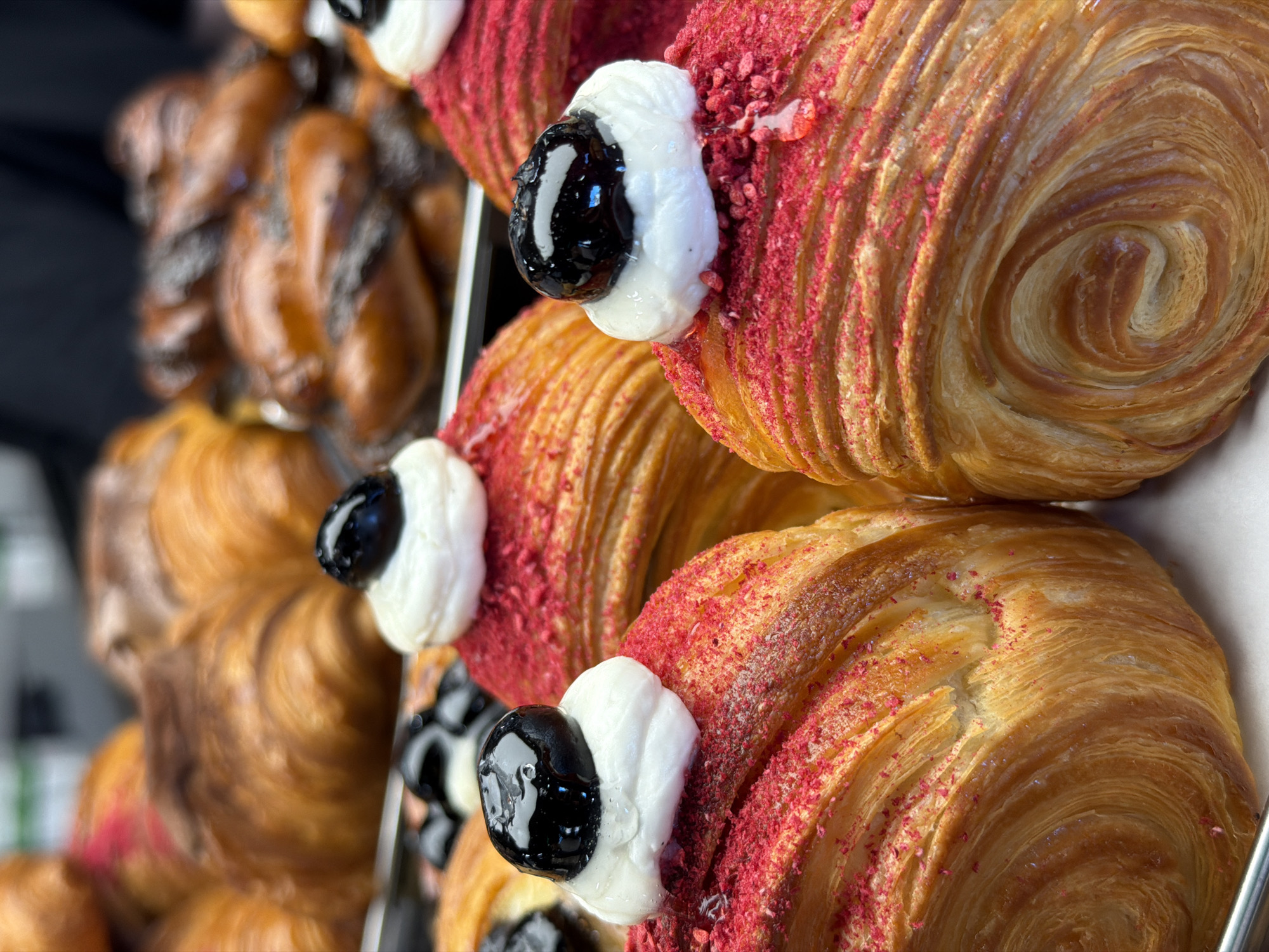 A close-up shot depicts a bakery display with various pastries. Three pain au chocolat croissants with red sprinkles are stacked on the right side, each topped with a dollop of white cream and a shiny, dark cherry. To the left, out of focus, there are other brown pastries. The location is unknown and there is no information about the city of Izhevsk, Russia.
