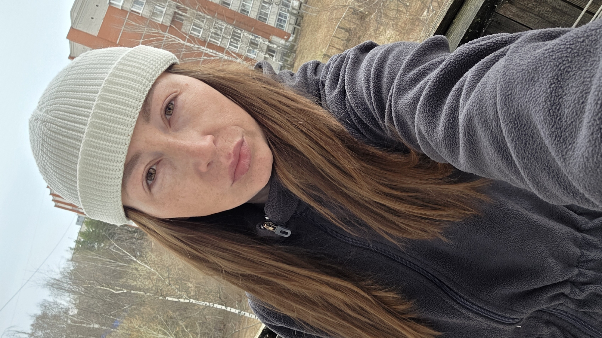 A woman with light brown hair and freckles is wearing a light-colored beanie and a dark gray fleece jacket with a zipper. The background shows a building with multiple windows and bare trees, suggesting it is an urban area in a temperate zone. The woman appears to be taking a selfie outdoors.