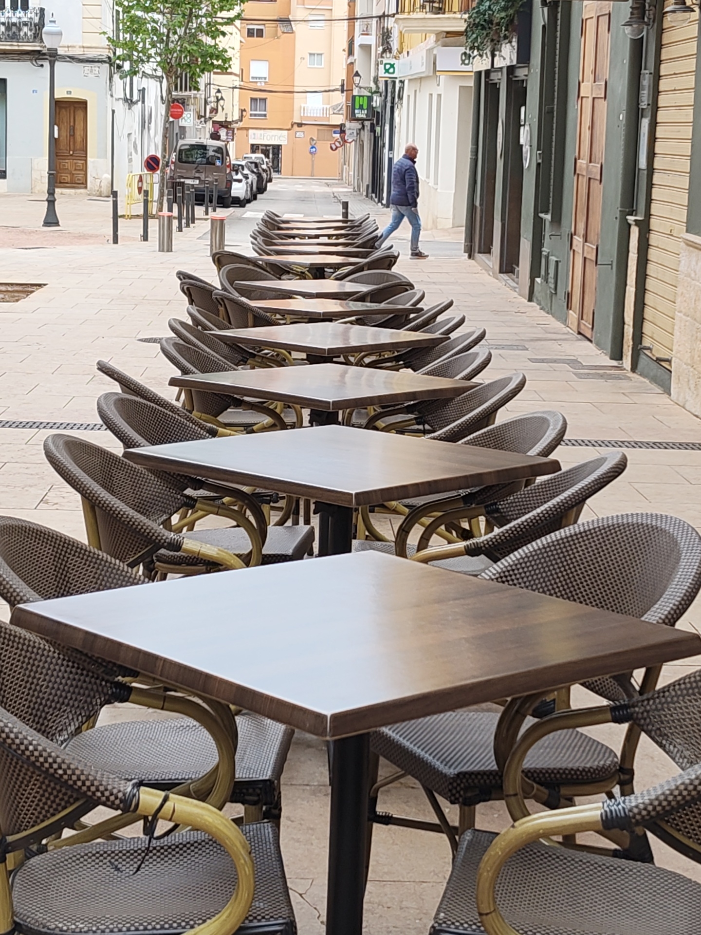 The image shows a row of square tables and chairs, seemingly part of an outdoor cafe seating arrangement, on a pedestrian street in Dénia, Spain. The tables are dark wood with black metal bases, and the chairs are woven rattan or a similar material, with curved arms. The pavement is made of light-colored rectangular tiles. In the background, a man in a blue jacket and jeans is walking away from the camera along the street. There are buildings on either side of the street, with various storefronts and architectural details. Parked cars are visible further down the street. The scene appears to be during daylight hours.