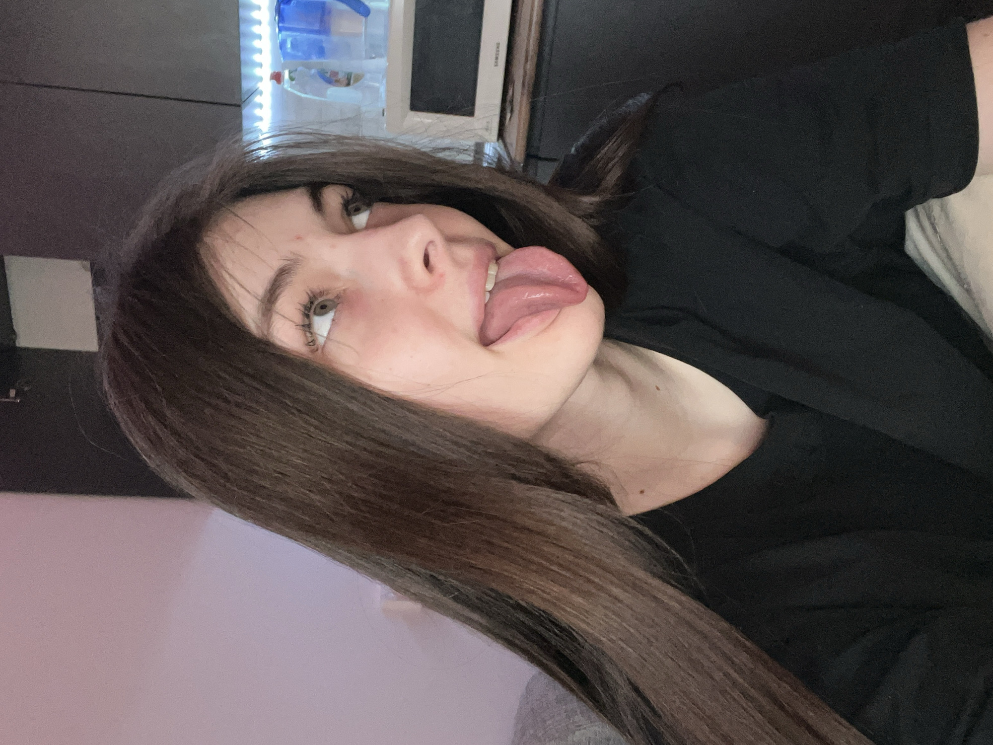 A young woman with long, brown hair is sticking out her tongue. She is wearing a black t-shirt. In the background, there is a television, as well as a light-colored wall. The location is not identifiable as Saint Petersburg, Russia.