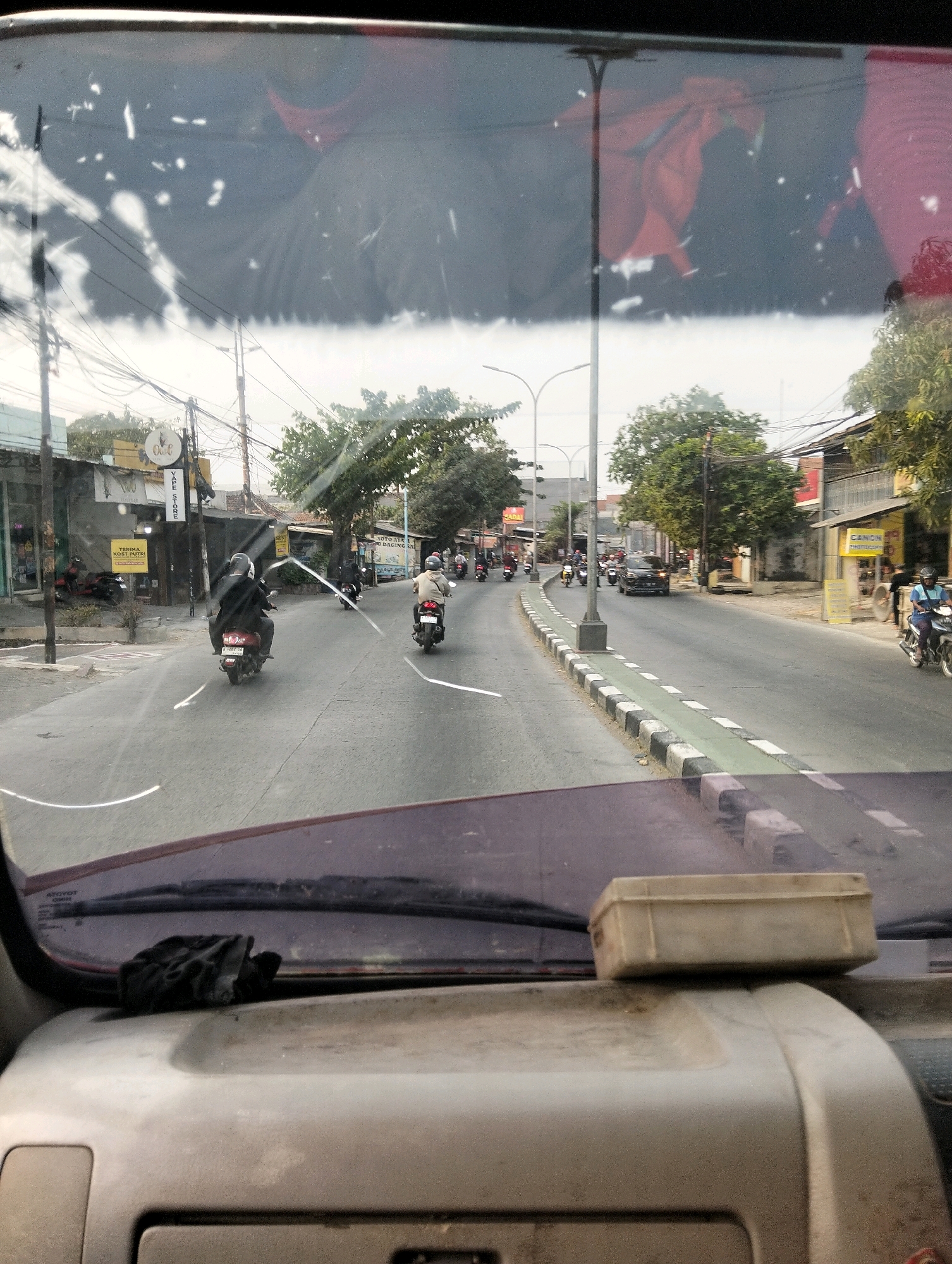 The image is taken from inside a vehicle, looking out through the windshield at a street scene in Semarang, Indonesia. The windshield is dirty, with several white smudges and streaks obscuring the view. In the foreground, the top of the vehicle's dashboard is visible, with a dusty surface and a small, light-colored rectangular box resting on it.
The street is paved and appears to be moderately busy. There are several motorcycles on the road, some with riders. One motorcycle in the middle distance has a rider wearing a dark jacket and a helmet. To the right, another motorcycle has a rider wearing a light-colored jacket and a helmet. Further down the road, a dark-colored car is visible, along with more motorcycles.
Buildings line both sides of the street. On the left side, there are several shops with signage, including one that says "YAE STORE." On the right side, more buildings and shops are present. Trees are interspersed along the street. Overhead power lines are visible. The overall lighting suggests it is daytime, possibly overcast. The presence of the Indonesian script on the signs confirms the location as Indonesia.