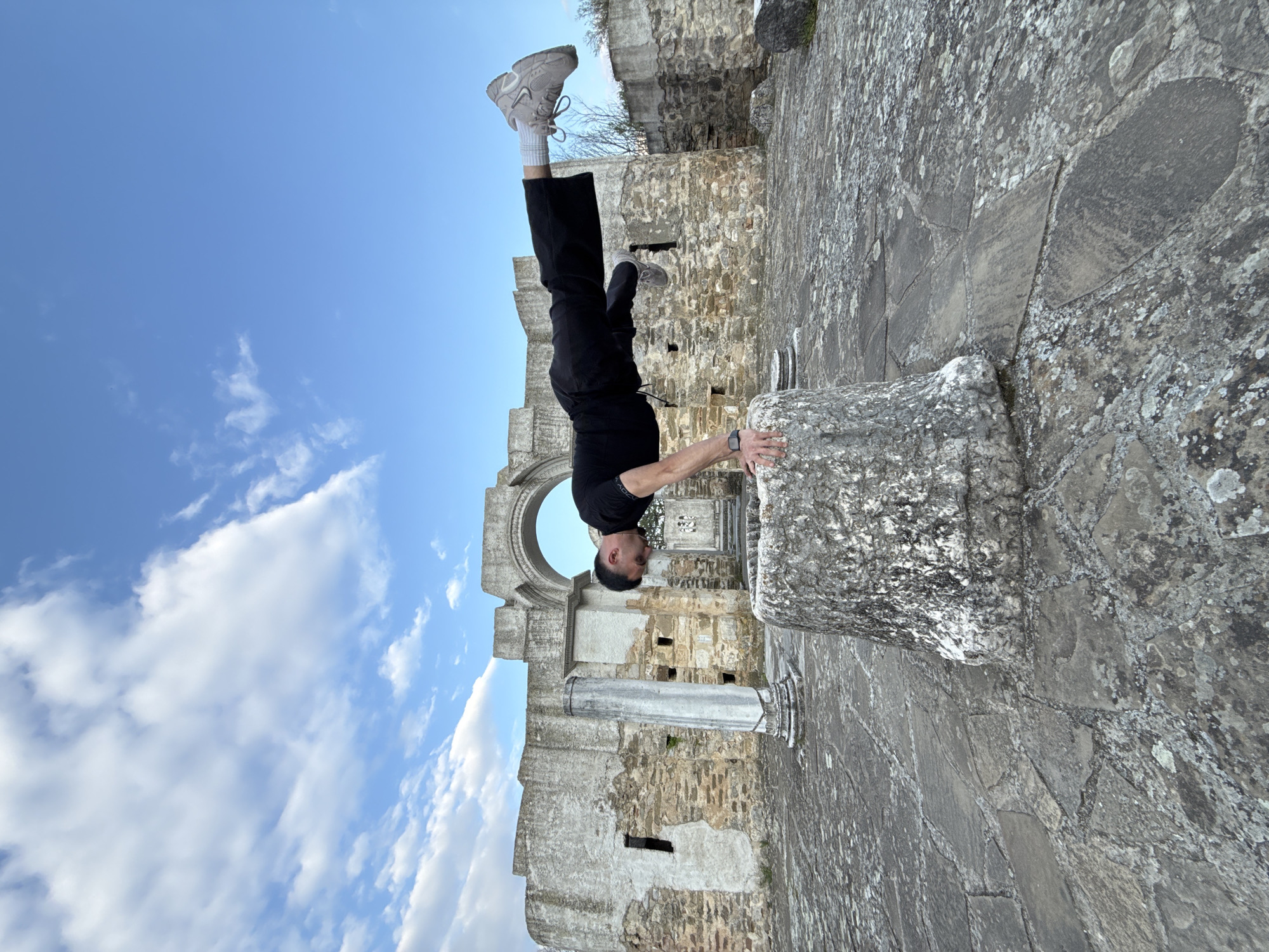 A man is performing a handstand on top of a stone pillar in what appears to be the ruins of a building. He is wearing a black shirt, black pants, white socks, and grey sneakers. His hands are on the edge of the pillar for balance. The ruins have stone walls and columns, with a large arched opening in the background. The sky above is blue with scattered white clouds. The ground around the pillar is made of large stone slabs.
The ruins are located in the city of Veliki Preslav, Bulgaria.