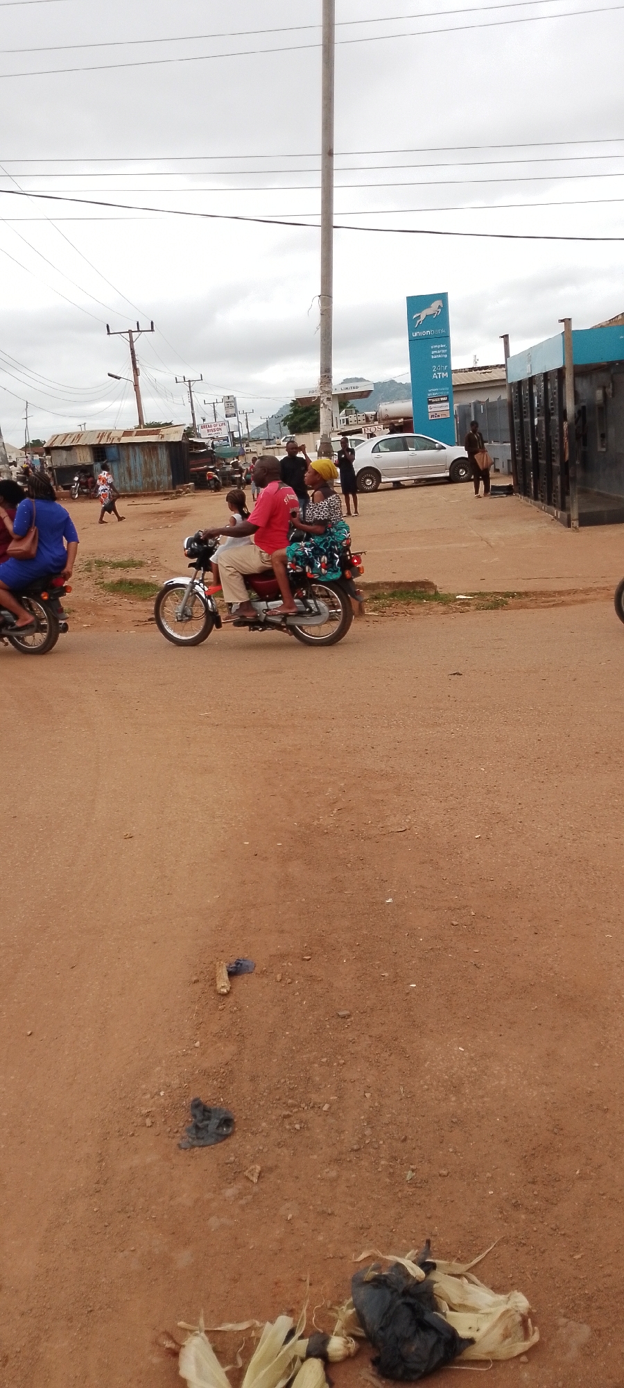 The image depicts a street scene in Gboko, Nigeria, likely during daylight hours with overcast skies. In the foreground, a dirt road is visible, with scattered debris including a piece of wood and some dark plastic. Further back, a man and two passengers, a woman and a child, are riding on a motorcycle. The man, wearing a red shirt and khaki pants, is driving, while the woman, in a patterned dress and yellow headscarf, sits behind him, holding onto him. A young child sits in front of the woman. To the left, another motorcycle with a rider in a blue dress is visible. In the background, a Union Bank ATM is prominently displayed on a building, along with other businesses. Several other individuals are seen walking or standing near the buildings and on the road. A silver car is parked near the ATM. Overhead, power lines and utility poles crisscross the sky. The environment appears to be a commercial or semi-urban area with corrugated metal structures and a mix of paved and unpaved surfaces.