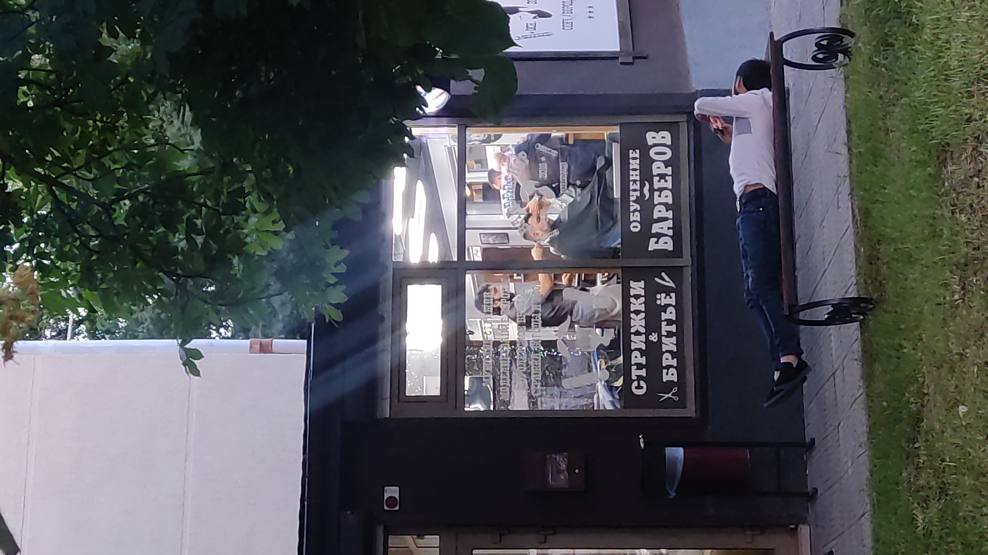 A man lies on a park bench in front of a barbershop, his head resting on his arm, eyes closed. He is wearing a white long-sleeved shirt and blue jeans. The barbershop's window displays the words "Стрижки & Бритьё" (Haircuts & Shaving) and "Обучение Барберов" (Barber Training). Inside, two barbers attend to customers, one holding scissors and comb, the other focused on a client's hair. The scene is set in the city of Krasnodar, Russia, evidenced by the Cyrillic signage. The lighting suggests it is late afternoon or early evening, with rays of sunlight piercing through the trees.