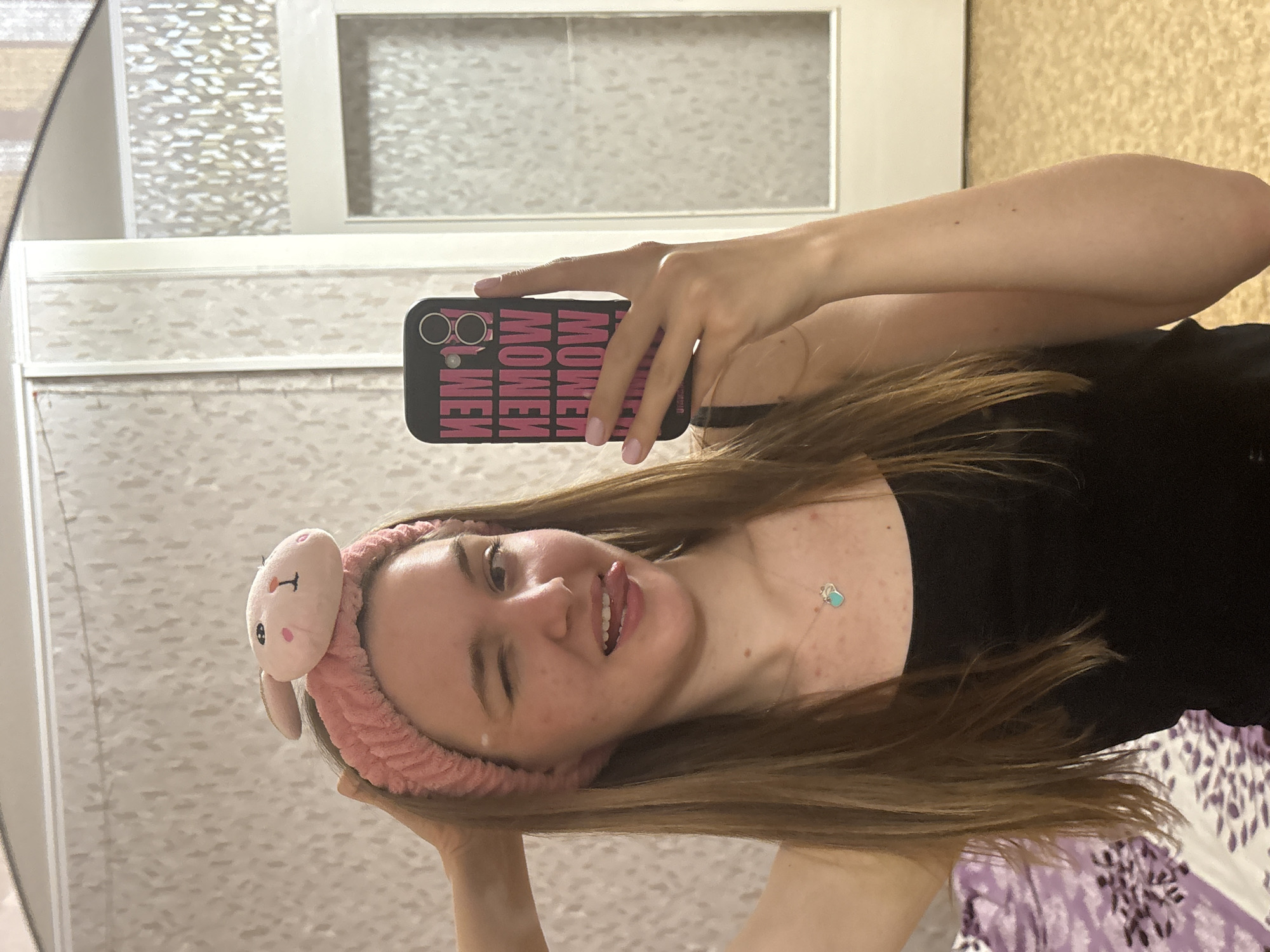 A selfie photograph taken indoors in Perm, Russia. A Caucasian woman with light brown hair wears a pink bunny-ear headband. She is winking and sticking out her tongue. She is holding a black smartphone with pink text on the back. She wears a black top. Behind her is a wall with textured beige and white wallpaper. A framed mirror is visible on the wall. A bed with a purple and white patterned duvet is partially visible at the bottom right.