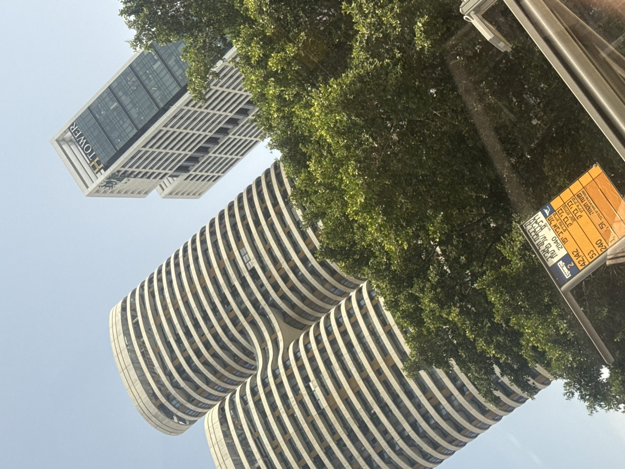 The image shows a view looking up at buildings in Tel Aviv, Israel. A building labeled "HTOWER" is in the upper left of the image, and a curved high-rise building is below it. In the foreground, there are tree branches and a bus stop sign with Hebrew text, likely near a station called Savidor Center. The bus stop sign displays information about bus numbers 42, 142, 53, and 240, along with their destinations like Bnei Brak, Petah Tikva, and Kiryat Shachidim.