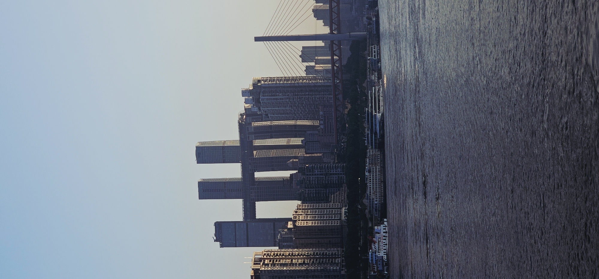 The image depicts a view of the Chongqing skyline in China, specifically the Nan'an District, from across a wide body of water. The foreground is dominated by the dark, rippling surface of the water, likely a river. In the middle ground, several large passenger boats are docked along the shoreline. Behind the boats, a dense collection of high-rise buildings forms the city's silhouette. A distinctive feature of the skyline is a pair of extremely tall, narrow skyscrapers that are connected by a horizontal structure resembling a bridge or walkway, creating an "H" shape. To the right of this structure, a cable-stayed bridge with a prominent pylon is visible, spanning the water and connecting to the cityscape. The sky is clear and a pale blue, suggesting it might be late afternoon or early morning. The overall impression is one of a modern, bustling metropolis situated along a major waterway. No people are discernible in the image.