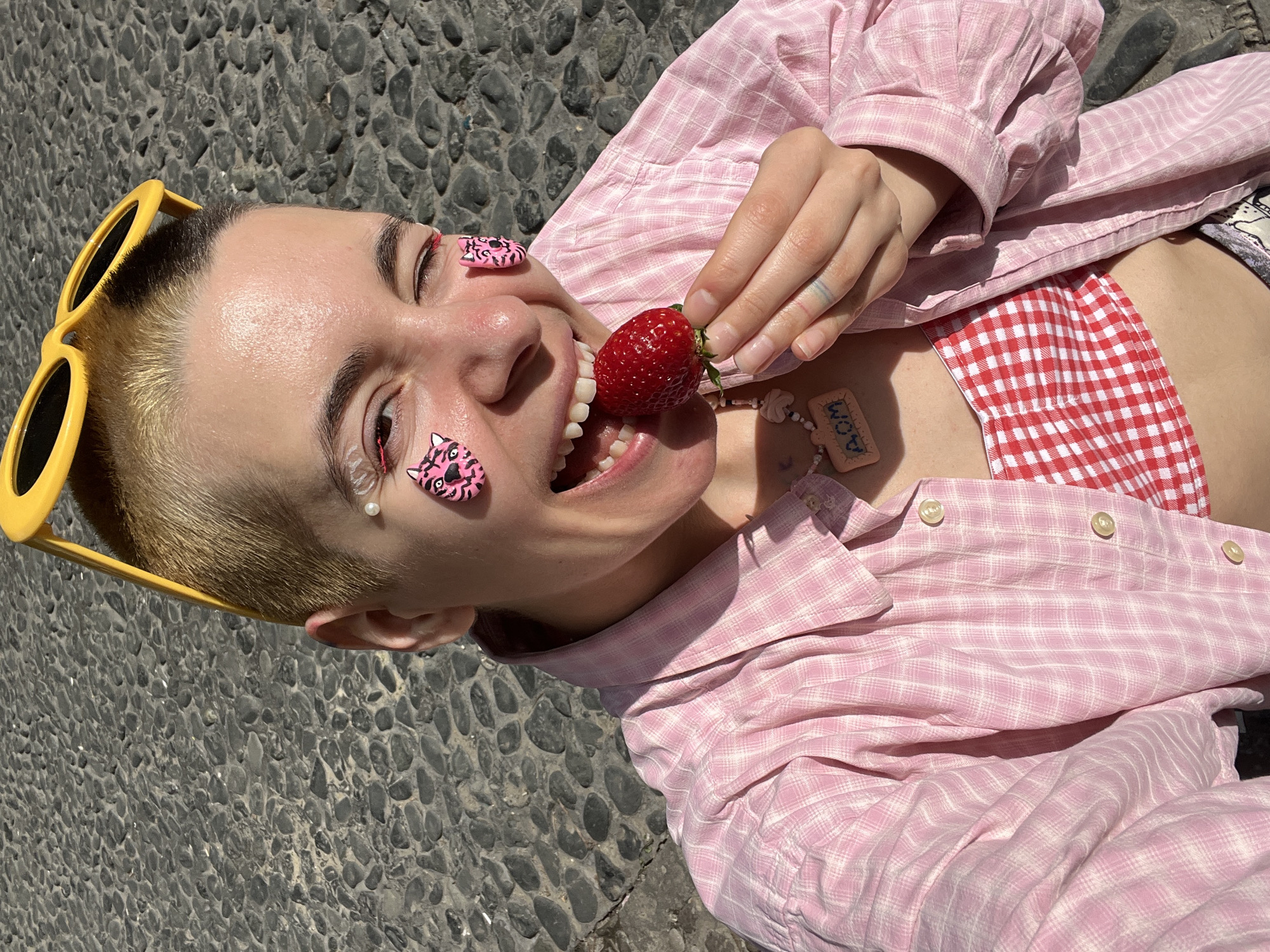 A person with short blonde hair and yellow sunglasses is lying on gray cobblestones, possibly in Belgrade, Serbia, although this cannot be confirmed from the image. They are wearing a pink and white plaid shirt over a red and white checkered top. The person has pink tiger stickers on their face, decorative pearls near their eye, and a "MOM" necklace. They are holding a strawberry to their mouth, appearing to be about to eat it. The background consists solely of the cobblestone pavement.