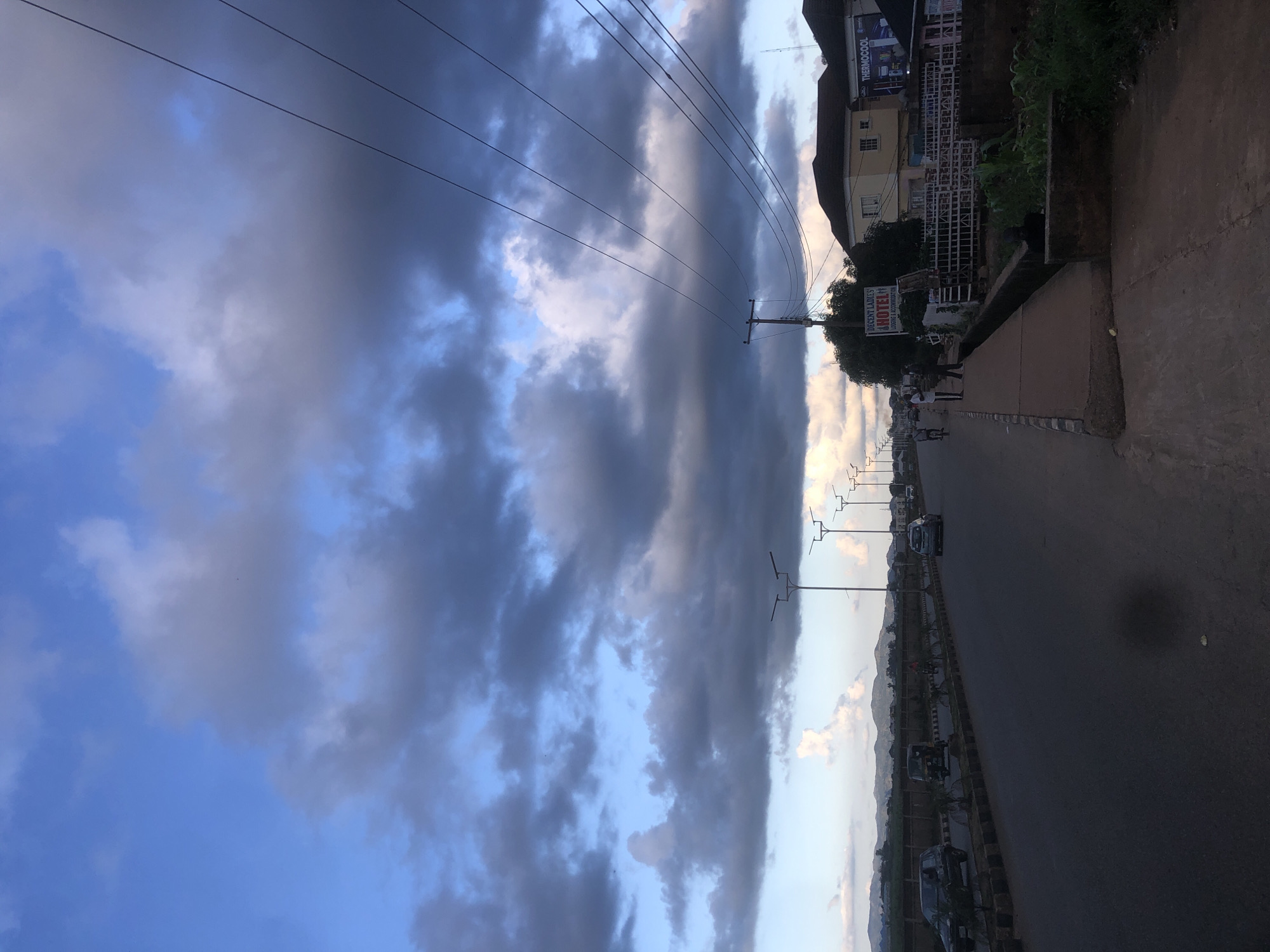 The image depicts a street scene in Barakin Lerawi, Nigeria, under a dramatic sky filled with dark, heavy clouds. A paved road runs horizontally across the frame, with vehicles including cars and a three-wheeled auto-rickshaw visible. A sidewalk runs along the right side of the road, bordered by a drainage ditch and vegetation. Buildings line the far side of the road, one of which displays a sign for "DECENT LADIES HOTEL." Electrical wires are strung overhead, supported by utility poles. The overall atmosphere suggests an impending change in weather, with the dark clouds casting a somber mood over the urban landscape.