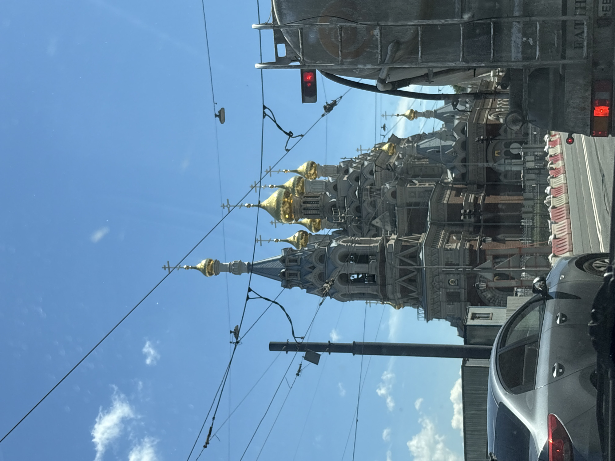 The image shows a view of a church with multiple gold-domed towers and intricate architectural details, resembling an Orthodox church in Saint Petersburg, Russia. The church is situated behind a street with traffic, including a silver car and the back of a large vehicle, possibly a truck, in the foreground. Overhead, a network of wires, likely for trolleybuses or trams, stretches across the sky. The sky is bright blue with scattered white clouds. The scene appears to be captured from inside a vehicle, looking out towards the street and the church. Construction barriers are visible along the side of the road. The overall impression is that of a city street scene with a prominent religious building.
