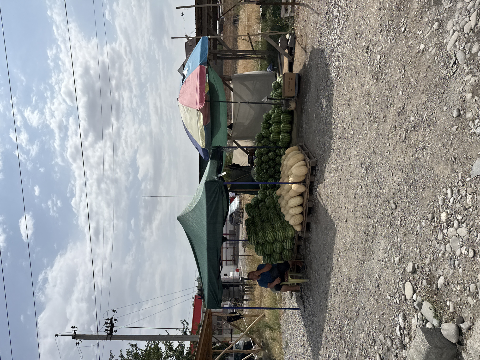 The image shows an outdoor market scene in Manas, Kyrgyzstan. A man is seated on a chair under a green canopy, seemingly resting or waiting for customers. In front of the canopy, a large pile of watermelons is displayed on wooden pallets. To the right of the watermelons, a pile of pale, elongated melons is also displayed on pallets. A multi-colored umbrella provides shade over a part of the market area. In the background, there are wooden structures, a fence, and some dry vegetation. Power lines and a utility pole are visible in the upper part of the image, along with a cloudy sky. The ground is covered with gravel and small stones. A few cars are partially visible in the background, suggesting the market is located near a road or thoroughfare.