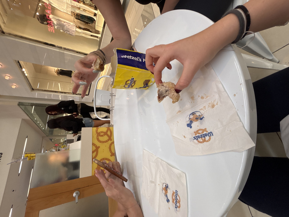 The image depicts an interior scene within a commercial shopping mall in Culver City, United States of America. In the immediate foreground, a white circular table is shown from a seated perspective. On the left side of the table, a person's hands are visible, holding and operating a light-colored smartphone. On the right side, another person's right hand holds a partially consumed, sugar-dusted pretzel bite over a "Wetzel's Pretzels" napkin that displays the brand logo and a Mickey Mouse pretzel graphic, alongside residual sugar and crumbs. A yellow "Wetzel's Pretzels" paper bag, containing additional pretzel bites, sits on the table between the two individuals.
In the mid-ground, a "Wetzel's Pretzels" counter is positioned. Its base features a yellow and brown pattern with pretzel motifs. Two women are standing at this counter. The woman on the left, with dark hair, wears a dark long-sleeved top and light denim shorts. The woman on the right, possessing reddish-brown hair, is dressed in a dark jacket, a leopard-print garment (possibly a top or accessory), and dark pants.
The background reveals a well-lit mall environment with white walls, recessed ceiling lighting, and reflective glass storefronts. One visible storefront clearly displays the word "GARAGE" and features mannequins dressed in various retail clothing items. A wooden door with a silver handle is partially visible to the left of the pretzel stand.