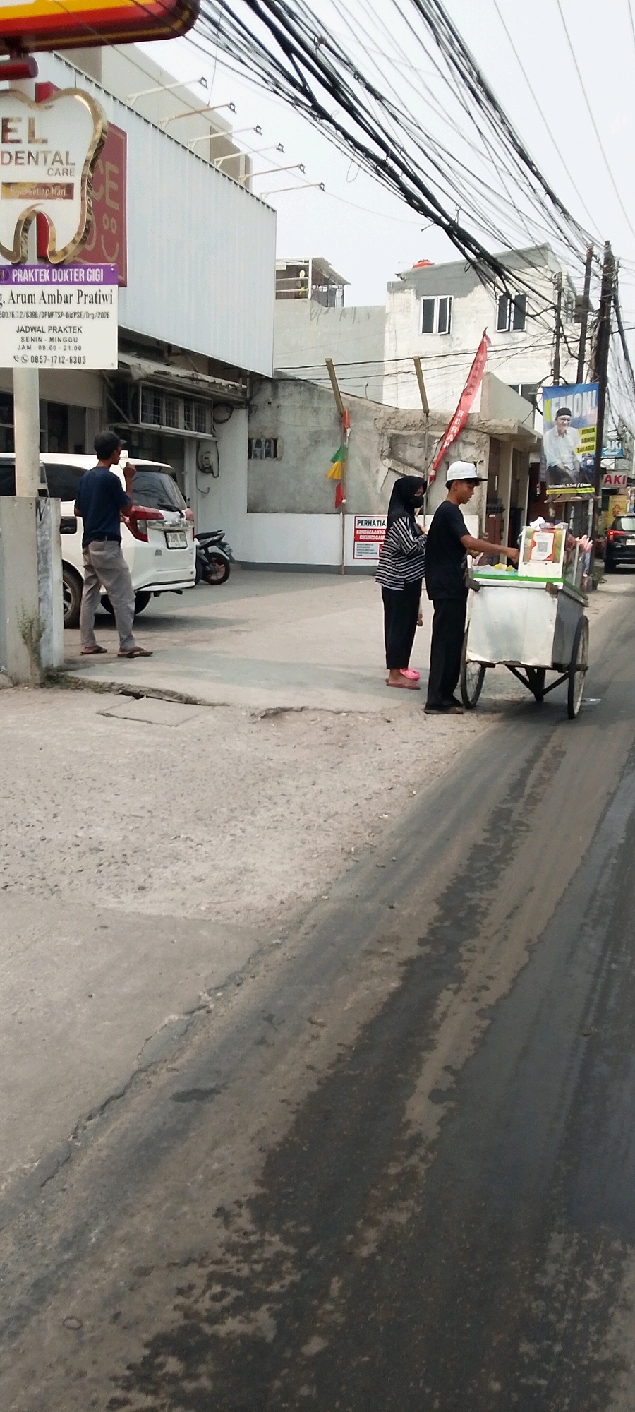 In Karangsatria, Indonesia, a man wearing a dark blue t-shirt and light grey pants stands near a white car, holding a drink. Beside him, a woman in a black and white striped shirt, black pants, and a black hijab stands next to a food cart. A man in a black t-shirt and a white cap operates the cart, which has a QR code displayed on it. The scene takes place on a street with a cracked asphalt surface. In the background, buildings line the street, including a dental clinic with a prominent tooth-shaped sign. Overhead, numerous electrical wires crisscross the sky.