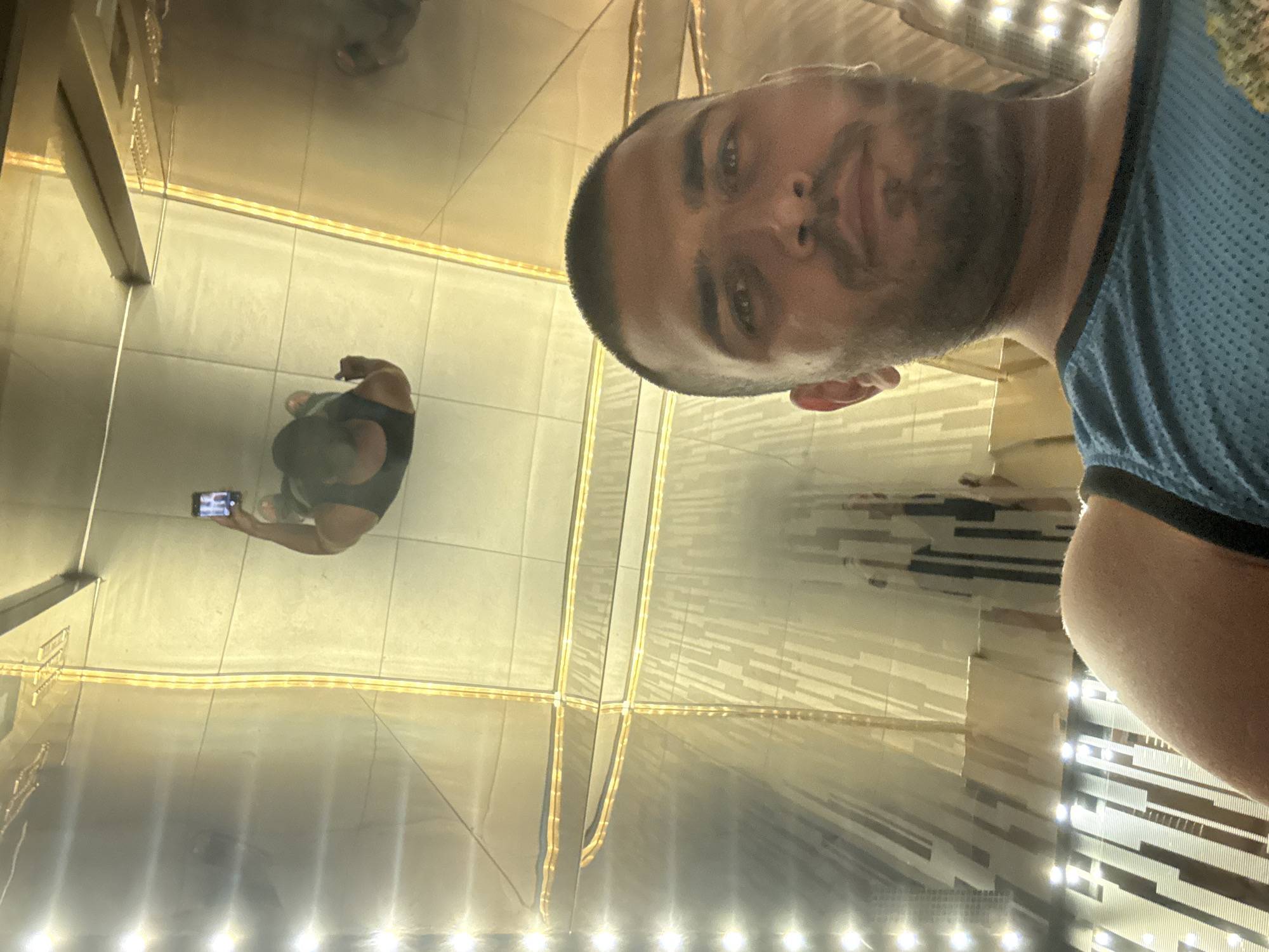 The image depicts a person taking a selfie in a mirror room. The person taking the selfie has short dark hair, a beard, and is wearing a blue tank top. Their reflection appears below, holding a phone. The room is illuminated by numerous lights, and the mirrored surfaces create multiple reflections. The location is likely the Petronas Twin Towers in Kuala Lumpur, Malaysia, based on the architectural style and the reflective nature of the surrounding walls. The person is interacting with the environment by taking a photo.