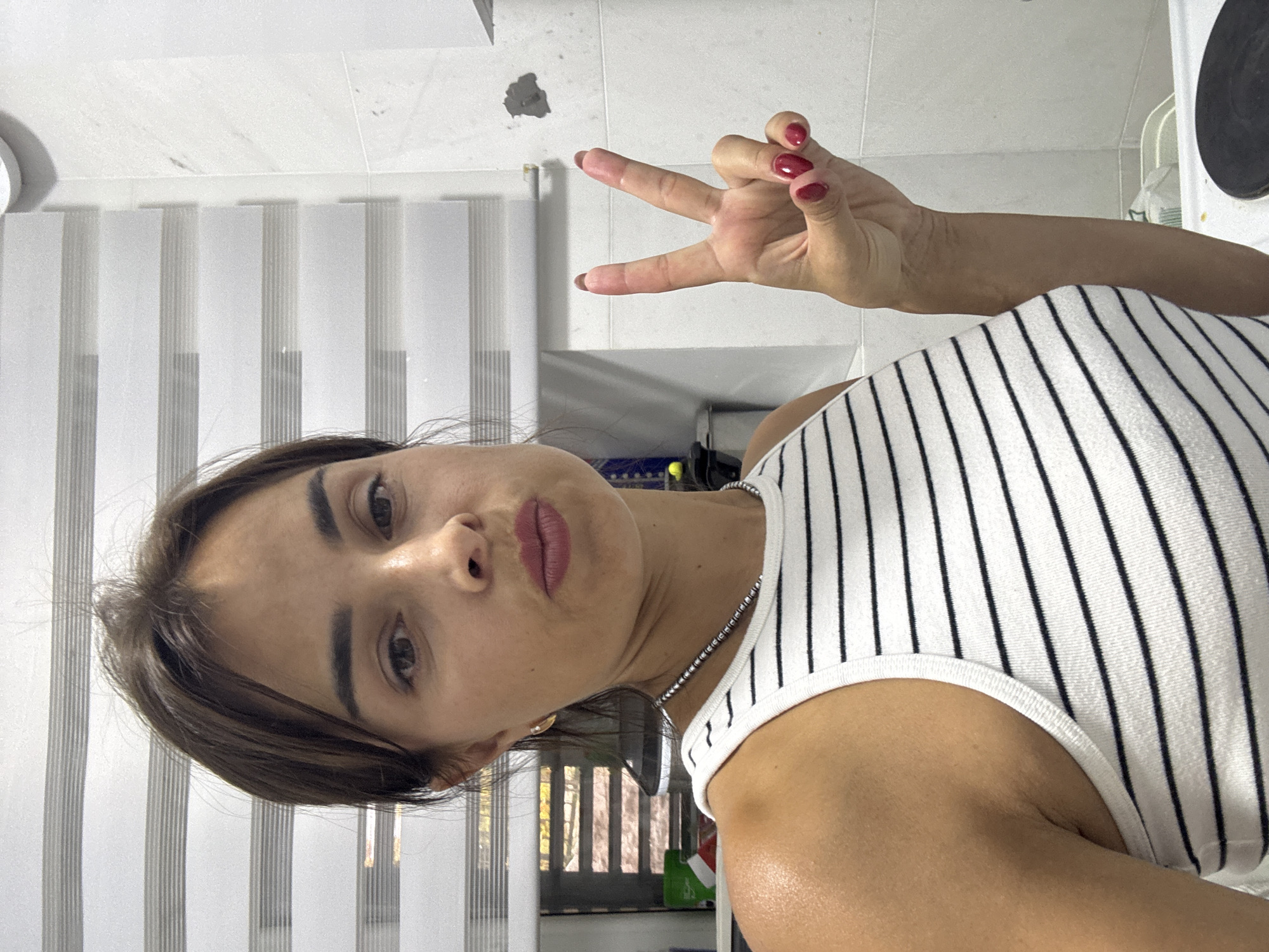 A young woman with dark hair pulled back is taking a selfie in what appears to be a bathroom or kitchen. She is wearing a white and black striped tank top and has on dark nail polish. She is holding up her right hand in a peace sign. The background consists of white blinds and tiled walls.
