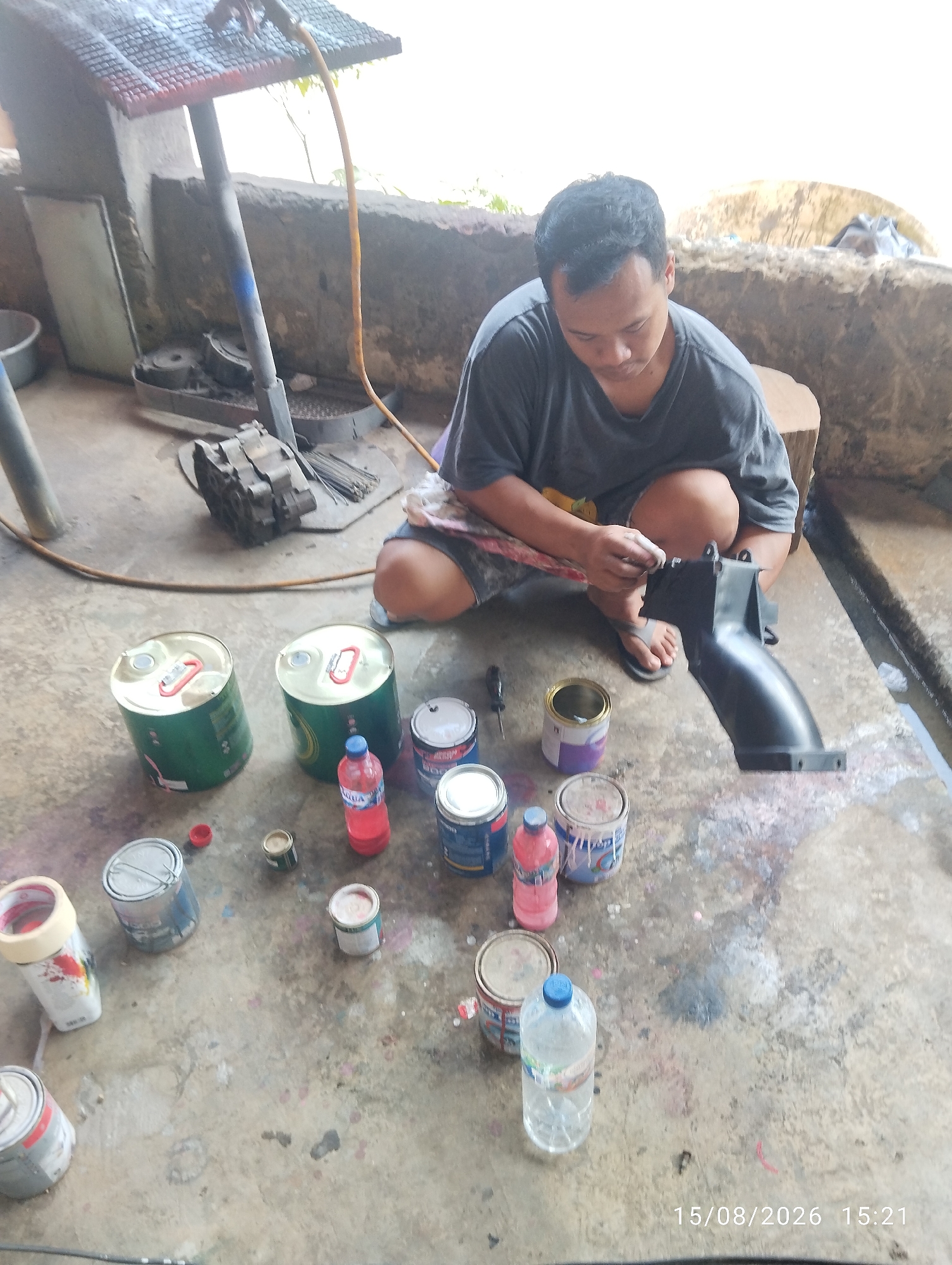 A man, wearing a dark gray t-shirt and plaid shorts, is sitting on the floor of an outdoor workshop in Tegal Rejo, Indonesia. He is in the process of painting a black, plastic automotive part. The floor is concrete and splattered with various colors of paint, indicating this is an active workspace. Around him are several open cans of paint, some still containing paint, and a red bottle of liquid, likely a solvent or thinner. There are also two large green metal cans, and a variety of smaller paint cans scattered on the ground. In the background, a metal stand with a grate on top is visible, along with what appears to be engine parts. The overall impression is of a practical, working environment where repairs or modifications are being made to vehicles.
