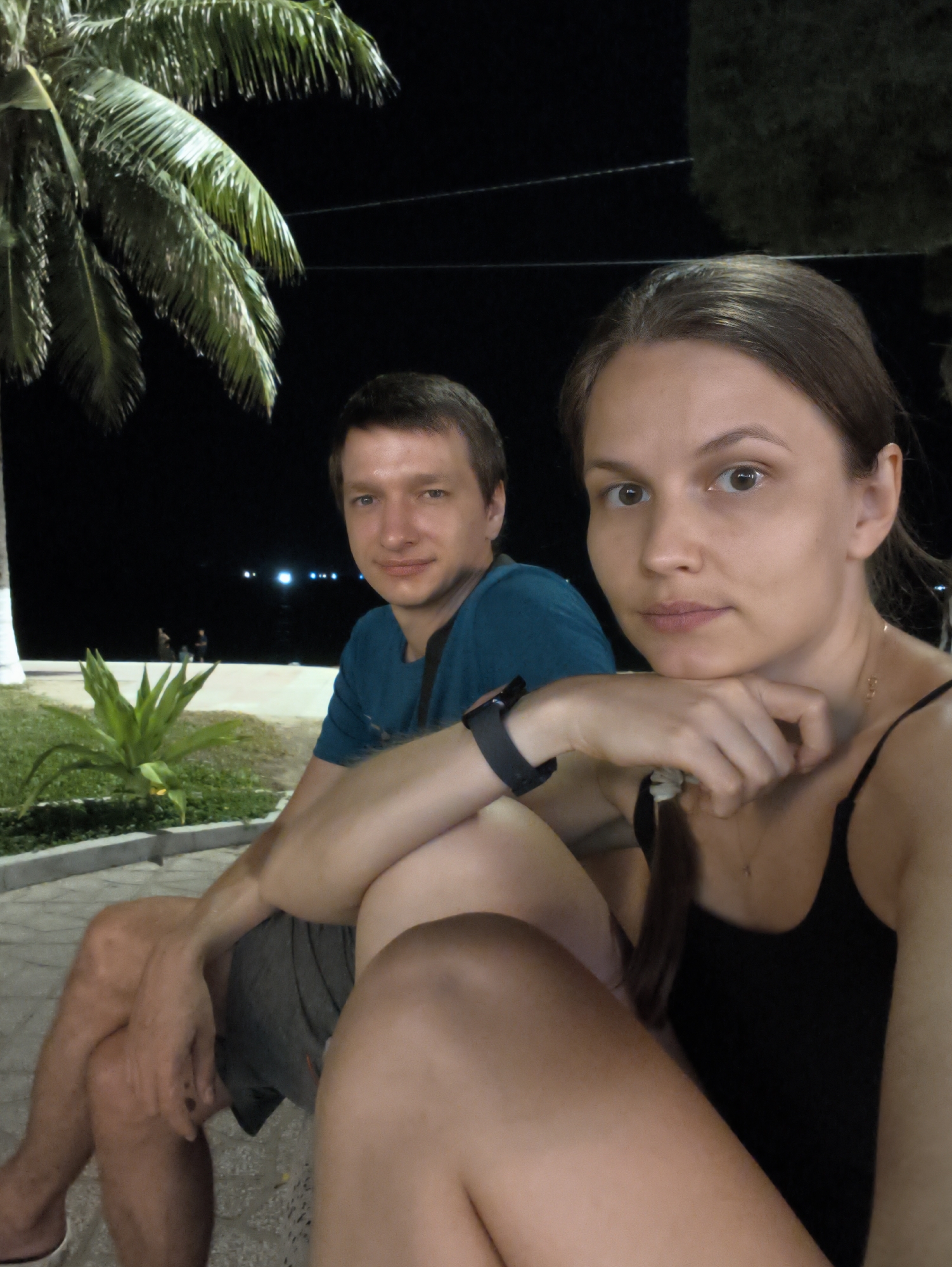 A man and a woman are sitting in front of a body of water at night, possibly in Nha Trang Ward, Vietnam, with a palm tree to the left and a plant in the foreground. The man is wearing a blue t-shirt and grey shorts. The woman is wearing a black tank top and has her hand on her chin. The background shows some lights from across the water.