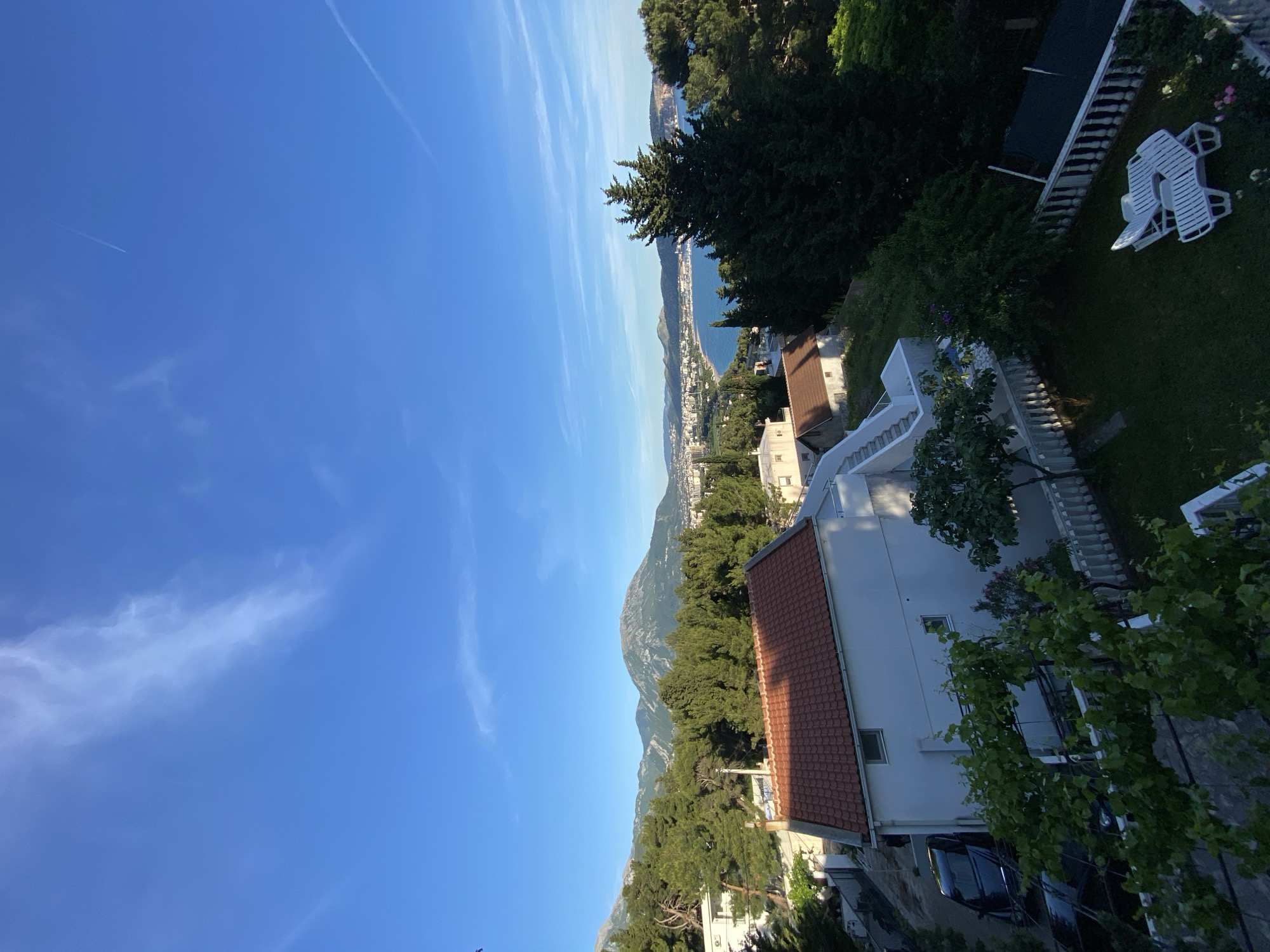 A view from an elevated position in Šušanj, Montenegro, reveals a landscape comprised of a clear blue sky with scattered clouds, a coastline with buildings, and a mountainous backdrop. In the foreground, there is a red-roofed building and a white building with multiple levels and a stairway. The surrounding vegetation includes trees and shrubbery. Lawn chairs and a ladder are visible in a grassy area. The scene is well-lit.