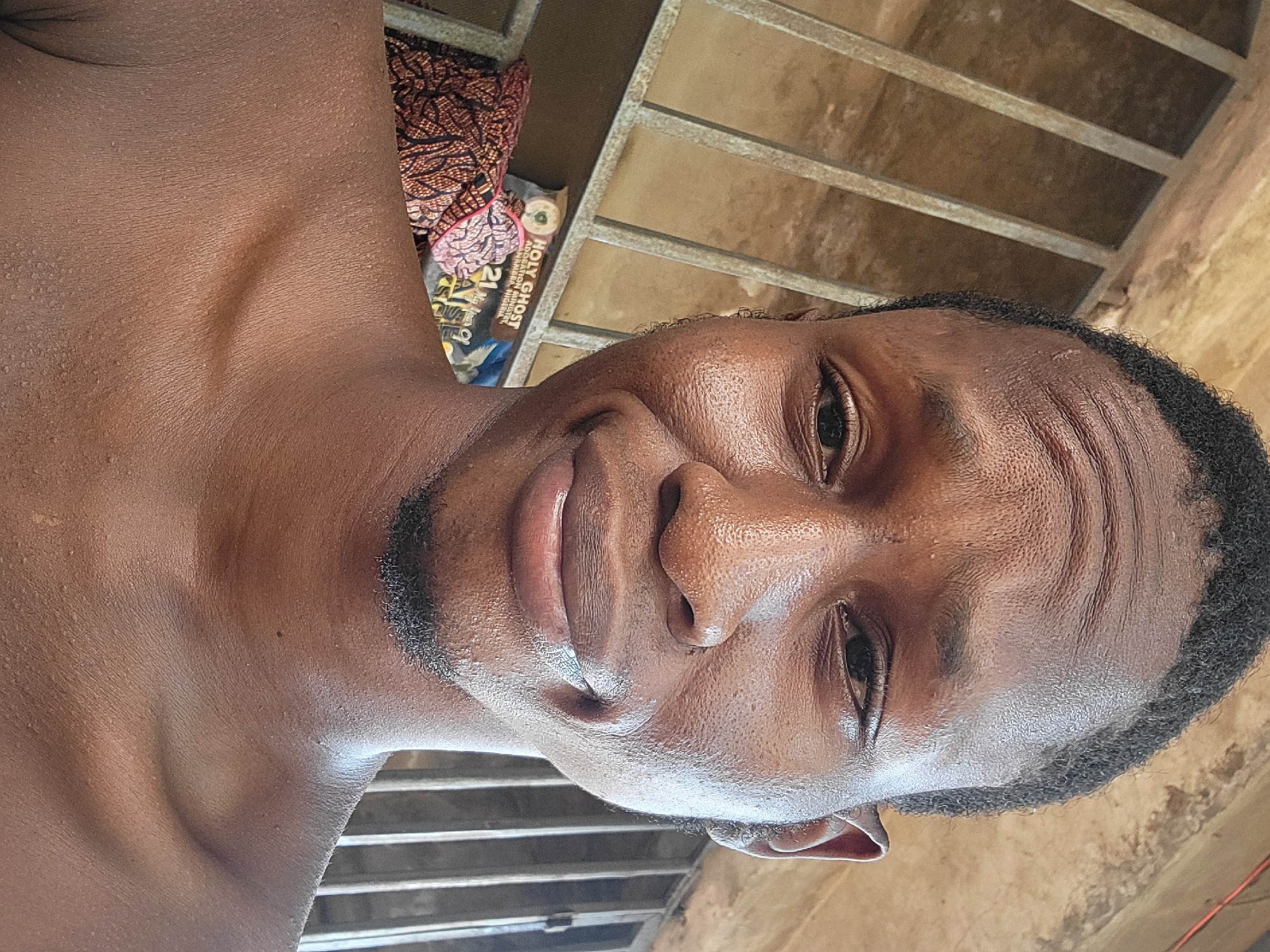 The image captures a close-up portrait of a young Black man, looking directly at the camera with a subtle, gentle smile. He has short dark hair, neatly trimmed stubble along his jawline and chin, and appears shirtless, revealing his upper chest and prominent collarbones. His skin has a slightly dewy or oily sheen, highlighting its texture and some faint lines around his eyes and on his forehead, which might suggest a warm climate.
In the background, behind his left shoulder, there is a glimpse of a colorful, patterned fabric, possibly an African wax print. Adjacent to this, a book or magazine cover is visible, clearly displaying the text "HOLY GHOST" at the top and "ADMINISTRATION NIGERIA" beneath it, indicating a religious publication originating from Nigeria. Above him and further into the background, a sturdy metal grate structure, resembling a window or ventilation cover, is seen against an earthy-brown, textured wall. The lighting suggests natural daylight, creating a soft illumination without harsh shadows.
The man's expression is relaxed and content, contributing to a calm mood. The details in the background, particularly the text "NIGERIA" and the architectural elements like the metal grate and textured wall, align with the provided context of the image being taken in Onitsha, Nigeria. The activity is simply posing for a photograph.