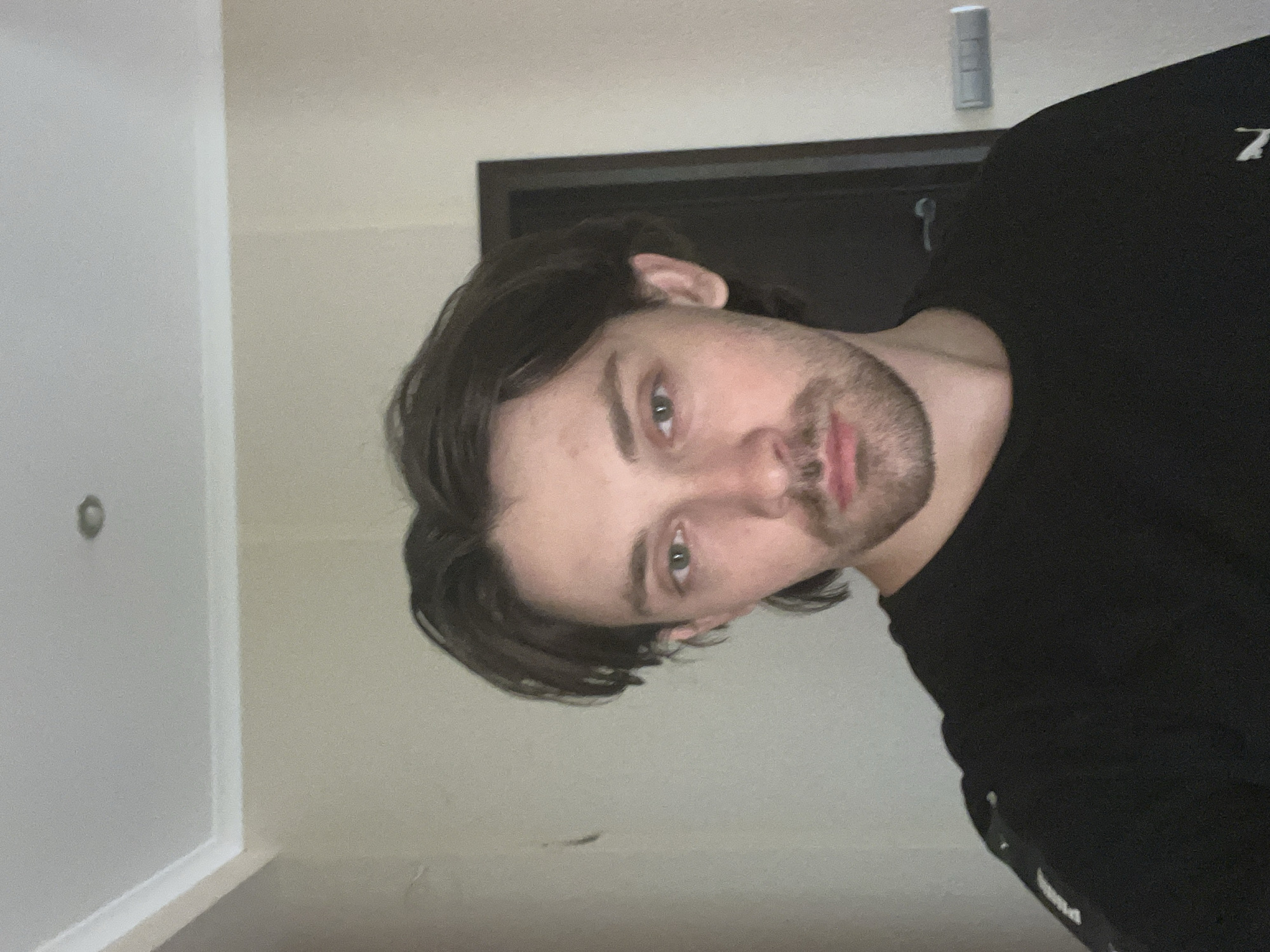 A headshot of a young man, rotated 90 degrees clockwise, is the central subject. He has medium-length dark brown hair, green eyes, and light stubble/a short beard. His expression is neutral as he looks directly at the camera. He is wearing a dark, likely black, long-sleeved shirt or sweatshirt, with a faint white symbol resembling the letter 'Z' visible on his right shoulder.
The setting is indoors, possibly a hallway or room in Ореховка, Russia. The background features light-colored walls: a cream or light beige wall to his right and a lighter, almost white wall to his left. A dark doorframe or door is partially visible behind his head and shoulder. On the wall above the door, there's a white rectangular object, potentially a light switch or thermostat. A circular object, possibly a light fixture or smoke detector, can be seen on the far left wall. The lighting suggests an indoor environment, either artificially lit or with indirect daylight. The image has a slightly grainy texture.