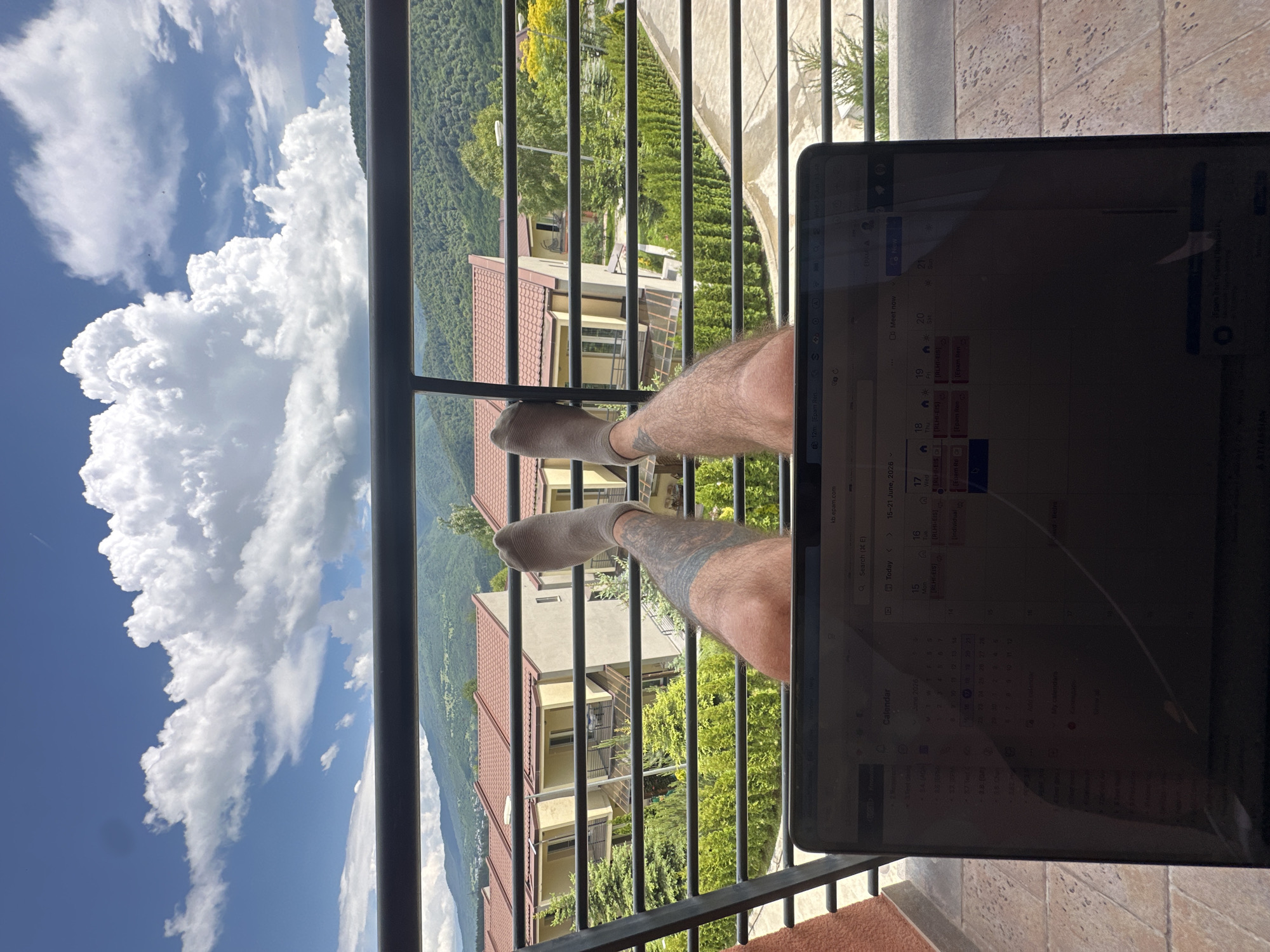 A first-person perspective shot from a balcony shows a person's legs propped up on the railing, wearing gray socks. The person's left leg has a prominent tattoo. In the foreground, a laptop is open on the balcony, displaying a calendar application. The background features a view of residential buildings with red roofs nestled amidst lush green hills under a bright blue sky with large, fluffy white clouds. The scene is set in Dilijan, Armenia, as indicated by the laptop's screen which shows "kb.epam.com" and other calendar entries, and implies a workspace setting with a scenic view.