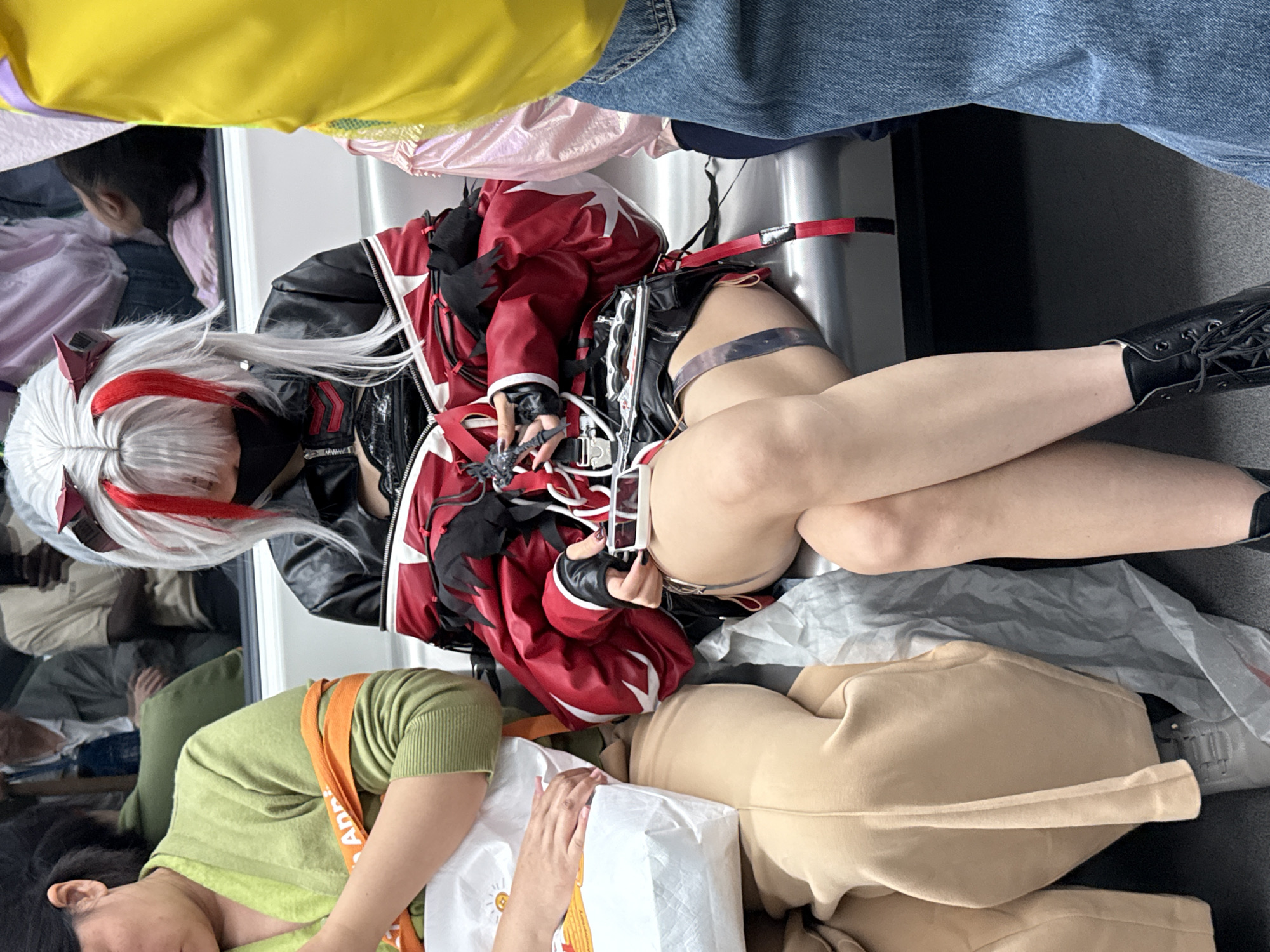 The image shows a gathering of people. One person is cosplaying with white hair and red accents, wearing a black and red outfit. Another person is wearing a green shirt with an orange strap and is holding a white shopping bag. Other individuals are present, with one wearing blue jeans and a yellow shirt. There is a gray plastic bag and tan clothing item near the cosplayer. The location appears to be indoors. The city is unknown.