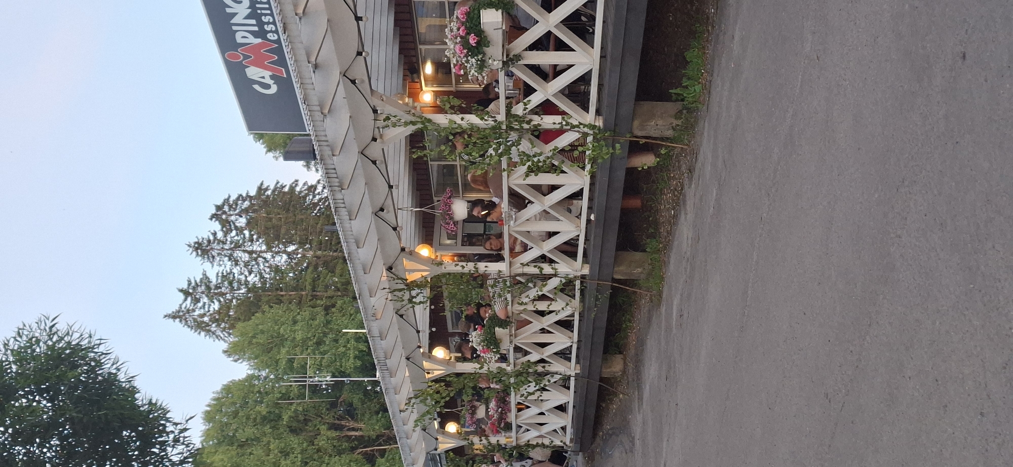 The image shows an outdoor seating area of a restaurant or cafe, identified by the sign "CAMPING Messilä" at the top right, indicating the location is Salpakangas, Finland. The area is covered by a white pergola decorated with hanging lights and climbing plants. White wooden railings with a criss-cross pattern line the elevated deck where patrons are seated at tables. Several people are visible, some eating and drinking, others engaged in conversation. Potted flowers, primarily pink and white, are placed along the railing and on tables, adding color to the scene. The background consists of green trees and a clear sky, suggesting a daytime or early evening setting. A paved area, likely a road or walkway, is in the foreground.