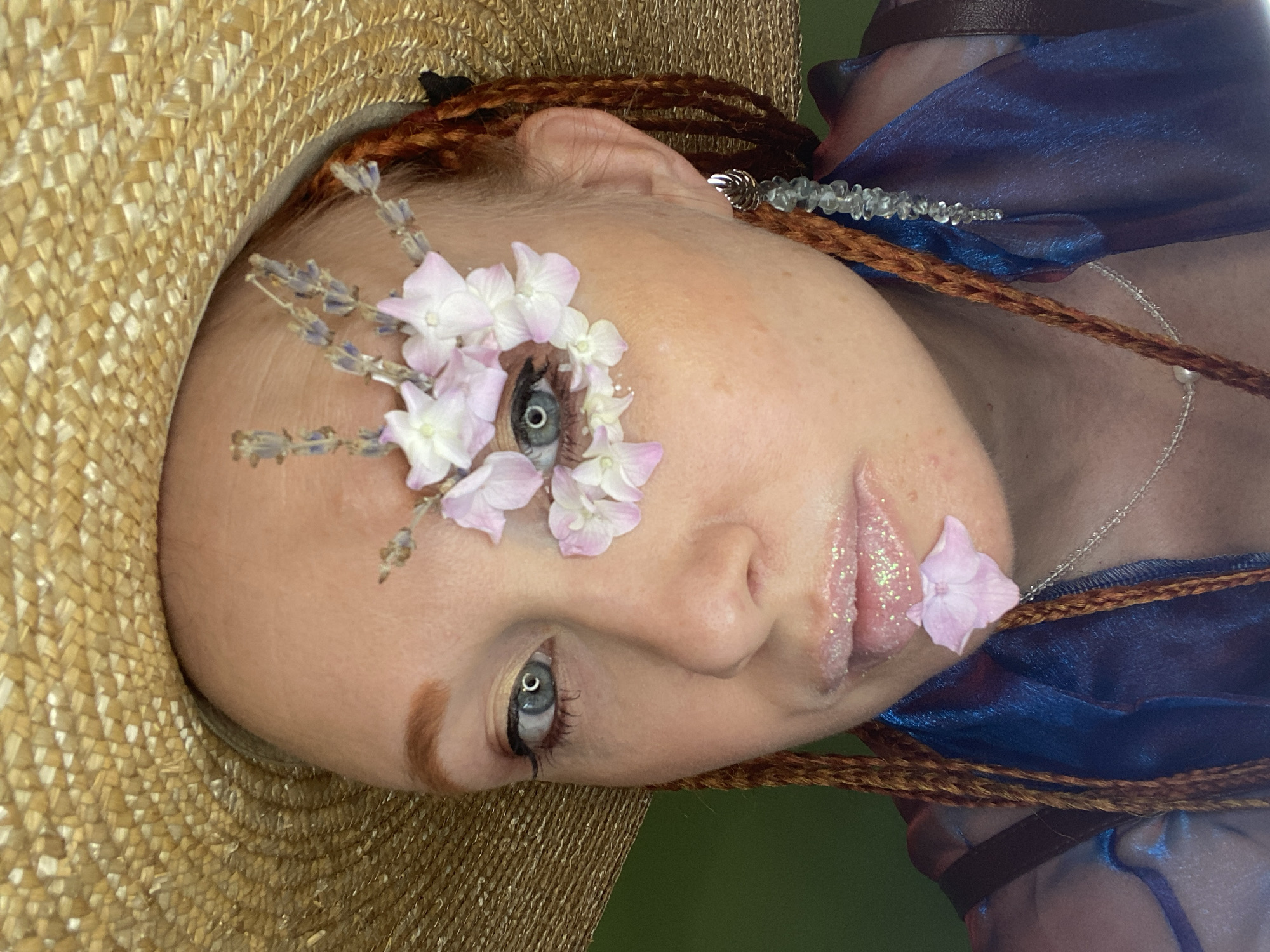 A close-up, front-facing, medium shot shows a person wearing a straw hat and an ornate makeup design. The person has fair skin, red hair styled in braids, and blue eyes. Their right eye is adorned with small pink and white flowers and lavender sprigs, with glitter accents. Their lips are glossy and also have glitter. A single pink flower petal rests on their lower lip. They are wearing a blue garment and a necklace. A light blue, beaded earring hangs from their right ear. The background is a solid green color. There is no information about the city of Sochi, Russia, in this image.