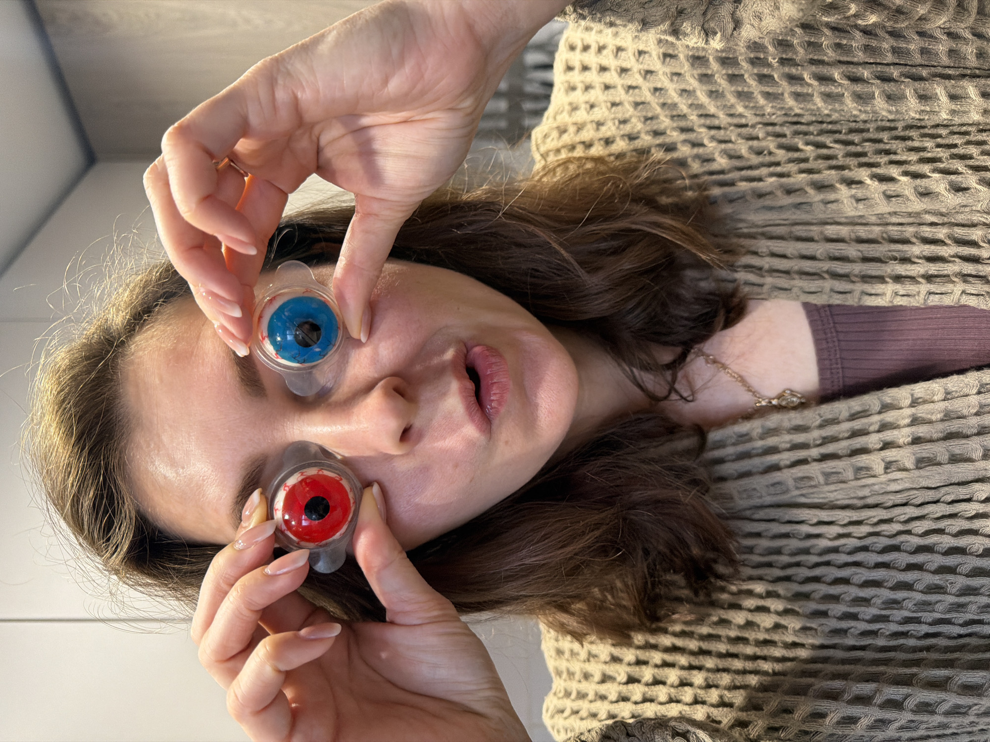 A woman is holding two artificial eyeballs in front of her face. The eyeball on the left is blue, and the eyeball on the right is red. She is wearing a brown sweater and a mauve-colored shirt underneath. Her hair is brown. The background is a plain white wall. The location is unknown and is not discernible from the image.