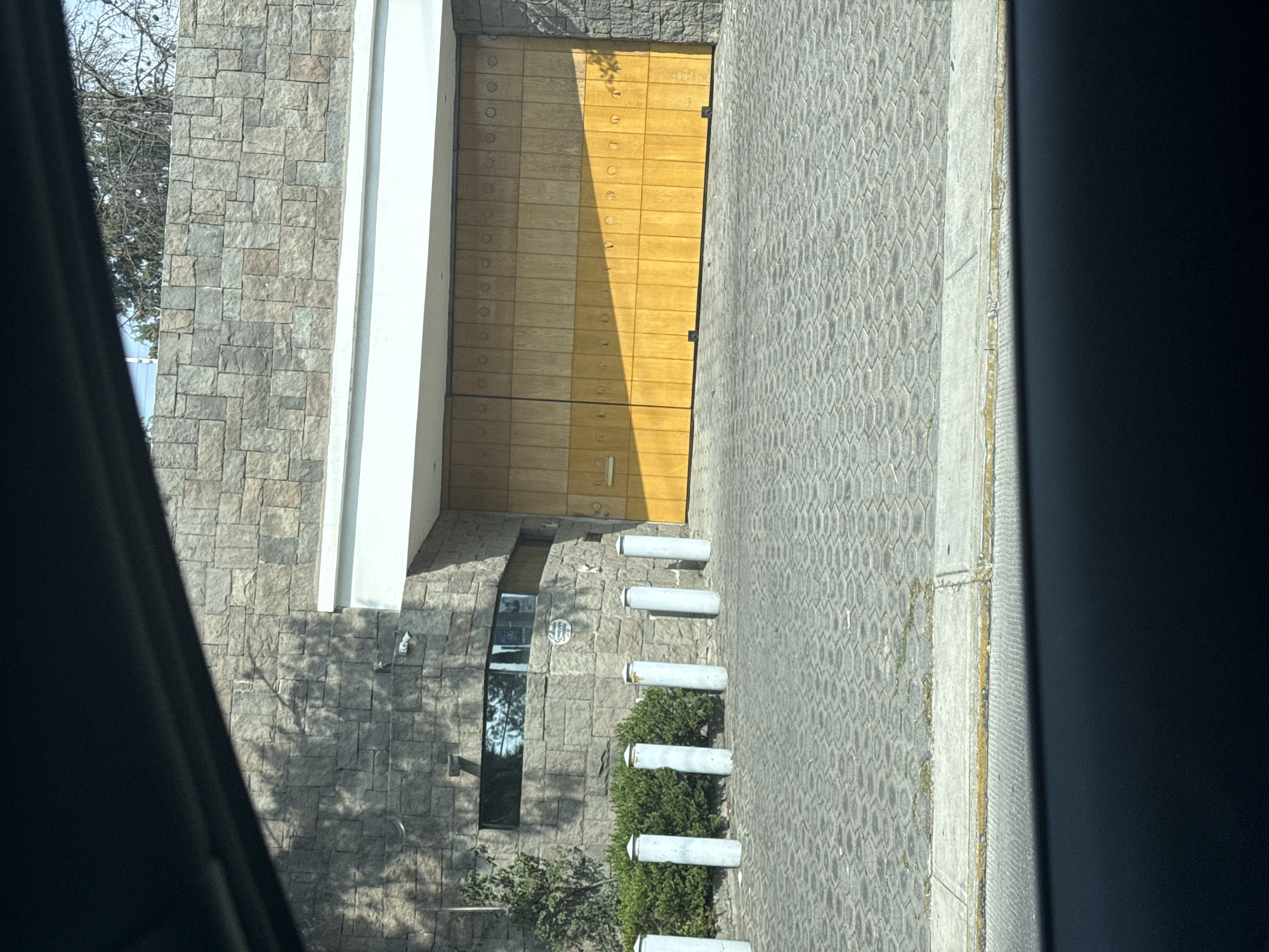 Here's a detailed description of the image:
**General Description:** The image shows the exterior of a building with a large wooden door and stone facade, possibly a residential or commercial structure. The perspective is from inside a vehicle, looking out towards the building and the adjacent paved area.
**Architectural Features:**
* **Facade:** The building is constructed with light-colored stone blocks, creating a textured surface.
* **Door:** The large wooden door is a prominent feature. It appears to be made of horizontal planks with metal detailing.
* **White Column:** There is a white column or decorative element alongside the door.
* **Window:** A tall, narrow window is visible to the left of the door.
* **Bolards:** There are a series of short, white cylindrical bollards placed along the edge of the building, likely to prevent vehicles from getting too close.
**Environment:**
* **Pavement:** The area in front of the building is paved with interlocking gray blocks.
* **Vegetation:** Small bushes or shrubs are planted near the base of the building, interspersed between the bollards.
* **Trees:** Bare trees can be seen to the side of the building.
**Vehicle Interior:**
* The edge of a vehicle's interior (possibly a door frame or pillar) is visible on the left and right sides of the image, indicating that the photo was taken from inside a car.
**Location:** The image is taken from the city of Puebla, Mexico.