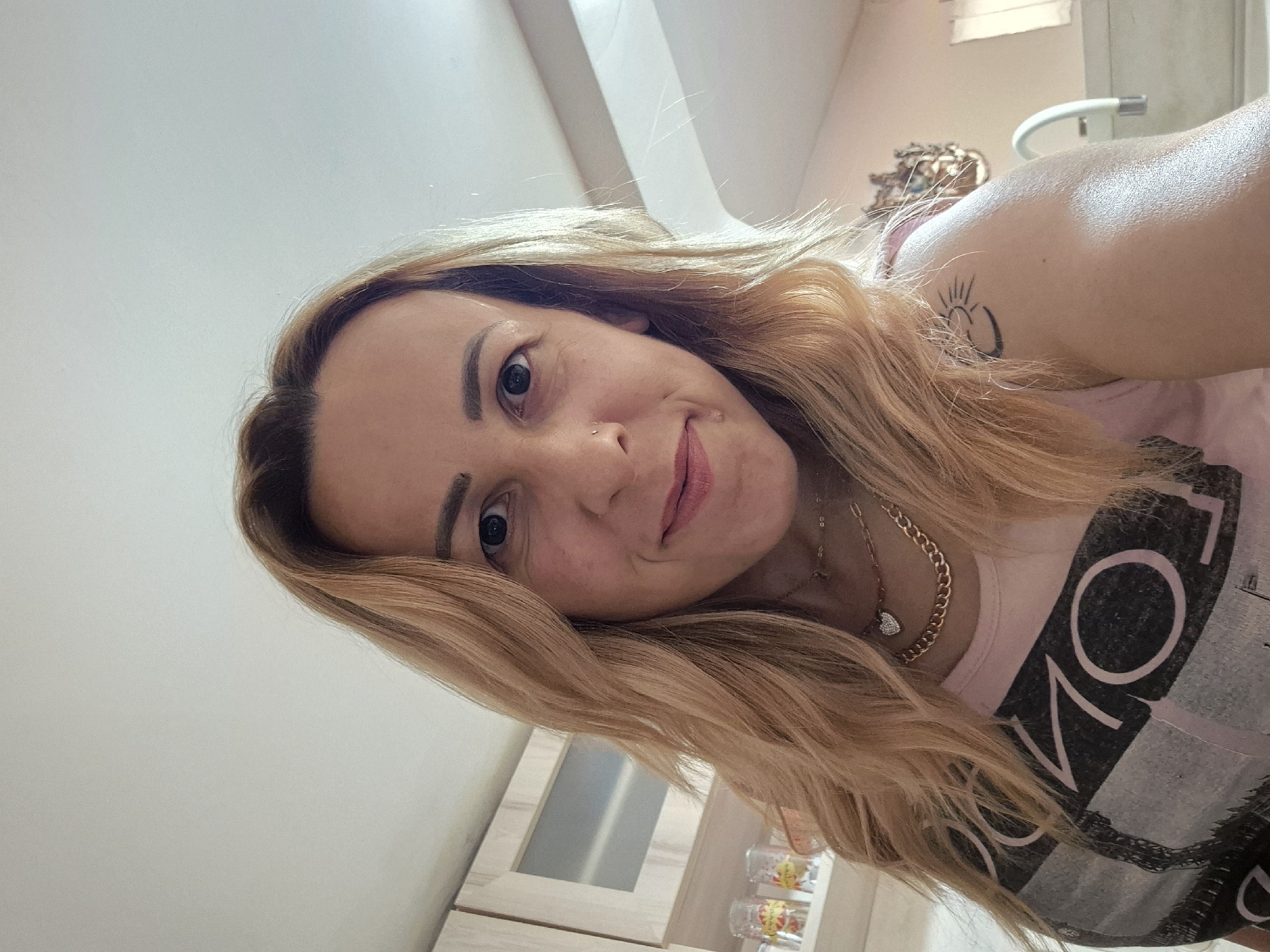 A medium shot captures a woman from the chest up, facing the camera. She has blonde hair that falls past her shoulders, framing a face with dark eyes and a slight smile. She is wearing a pink t-shirt with a graphic print of the word "LONDON". Around her neck, she wears two gold necklaces, one a chunky chain and the other a thinner chain with a small heart pendant. A tattoo of a sun and crescent moon is visible on her left shoulder. The background suggests an indoor setting, possibly a kitchen or living area, with cabinets and shelves visible. The lighting is bright, coming from overhead. The image appears to be a selfie.