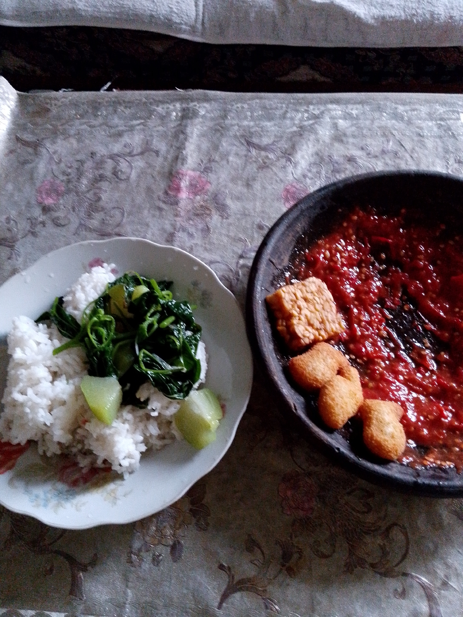 A plate of white rice topped with cooked green vegetables and sliced white vegetables sits on a patterned tablecloth next to a dark, round dish containing red chili paste with pieces of fried tempeh and what appear to be small fried fritters. The setting is likely a home in Babadan, Indonesia. The image is a top-down view of the meal.