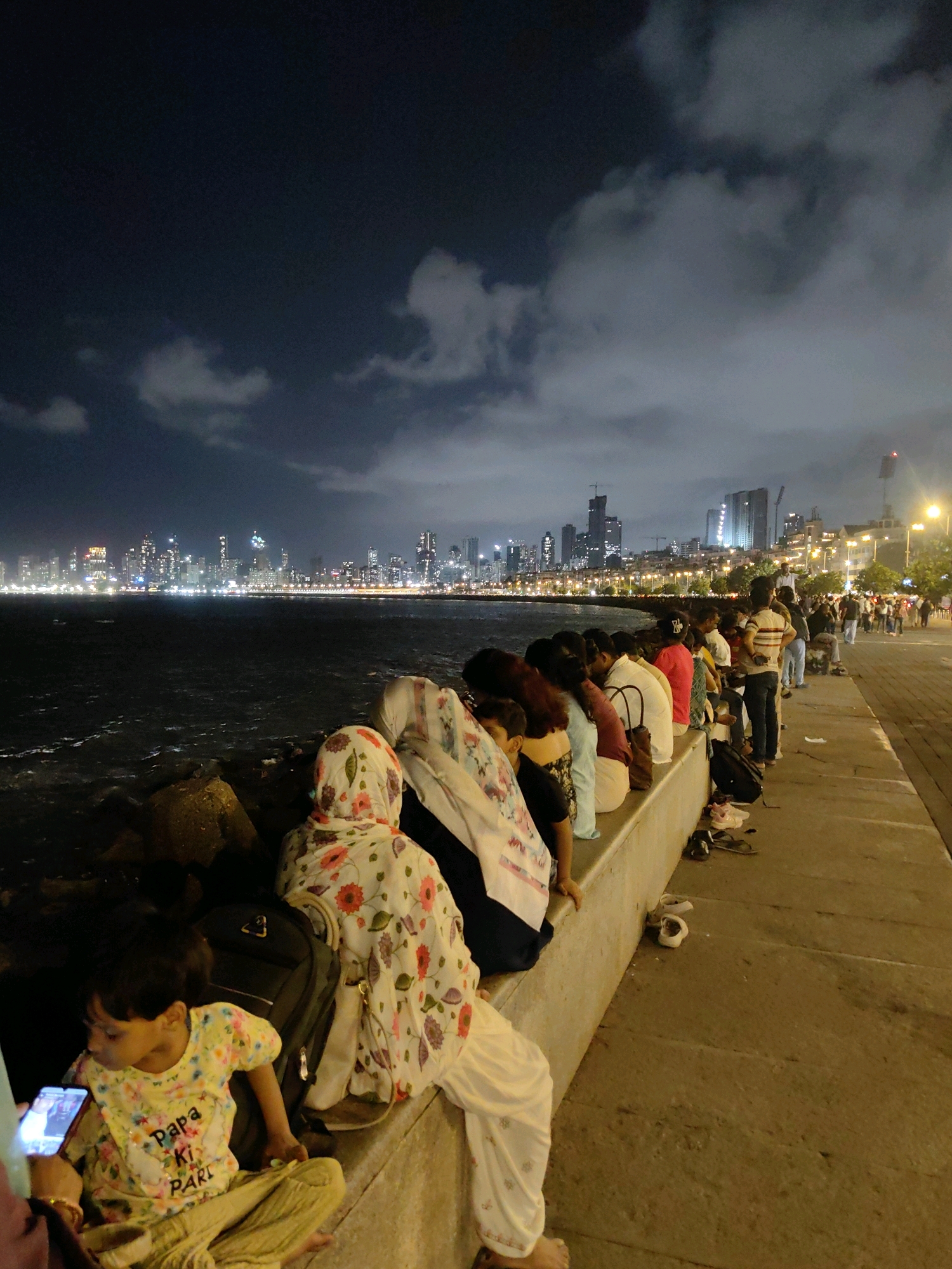 The image depicts a nighttime scene at Marine Drive in Mumbai, India, a popular promenade lined with a seawall. A group of people are seated along the seawall, facing the dark water and the distant, illuminated city skyline. The cityscape is characterized by numerous tall buildings with lit windows, creating a vibrant and bustling atmosphere. The sky is dark with some clouds, and streetlights illuminate the walkway beside the seawall. In the foreground, a young child is sitting on the ground, looking at a smartphone. The child is wearing a colorful t-shirt. Several pairs of footwear are scattered on the ground near the seawall. The overall mood of the scene is one of leisure and observation, with people enjoying the evening ambiance of the city.