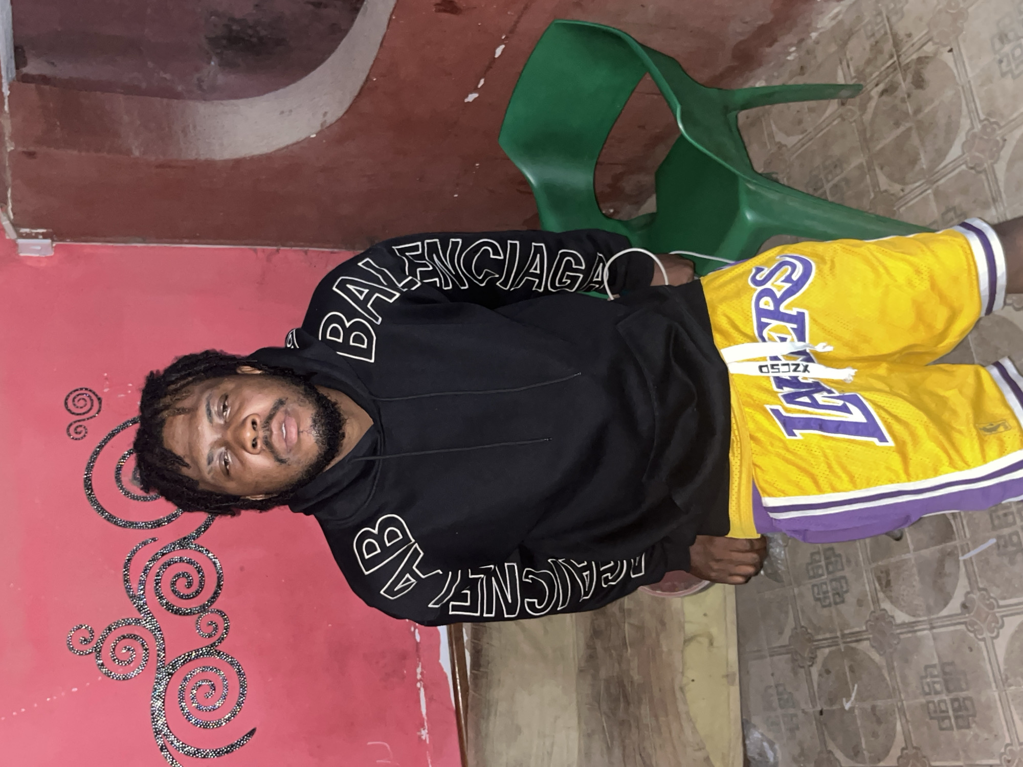 A young man with dreadlocks and a beard stands facing the camera in a room. He is wearing a black hoodie with white lettering on the sleeves that reads "BALENCIAGA" vertically, and yellow basketball shorts with purple and white trim, featuring "LAKERS" in large purple letters. A white drawstring hangs from the shorts. Behind him, the wall is painted a vibrant pink with black, glittery swirl patterns. To the right, a green plastic chair is visible. The floor is tiled with a brown and beige geometric pattern. The lighting appears to be artificial and the environment seems to be indoors, possibly in a residential or casual setting within Jos, Nigeria.