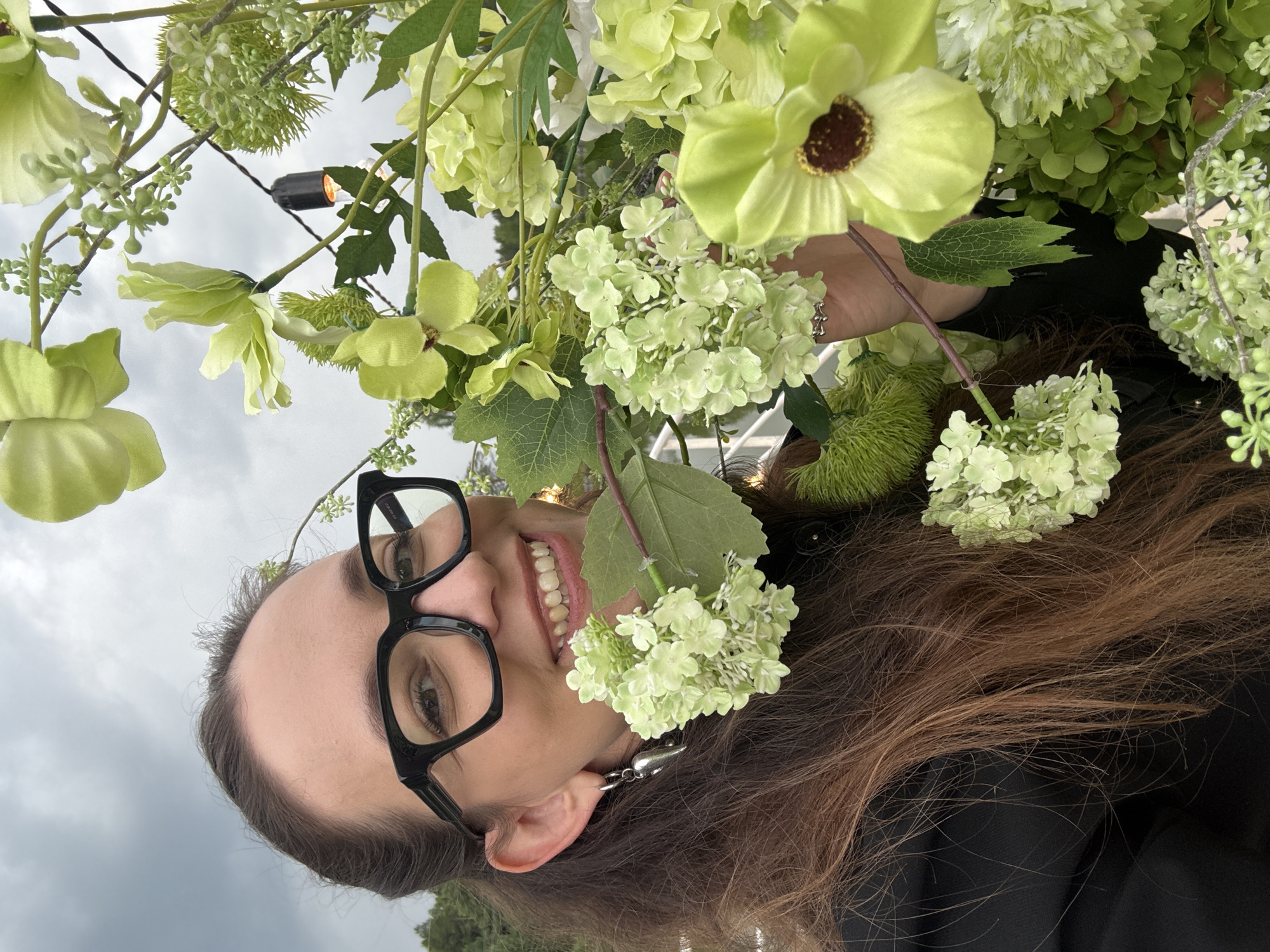 The image features a person with brown hair and glasses, smiling. They are positioned among an abundance of green flowers and foliage, with some string lights visible in the background. The person's face is turned towards the camera, and they are wearing large, black-rimmed glasses. The setting appears to be outdoors, possibly an event or decoration with a floral theme. The sky is overcast, suggesting a cloudy day. The person's expression conveys happiness and amusement. No text is visible in the image.
