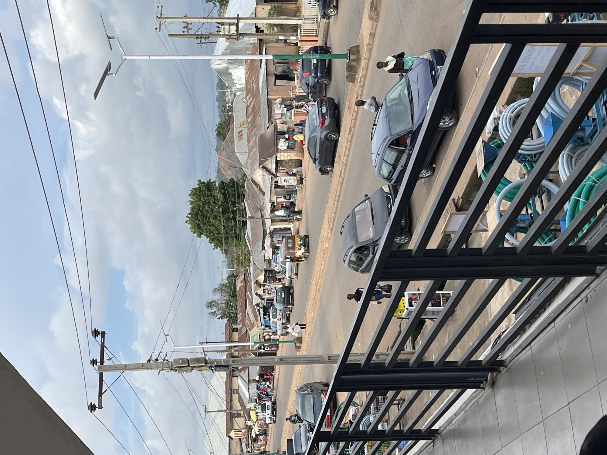 The image shows a street scene in Bukuru, Nigeria. The street is lined with buildings that appear to be shops or market stalls, with corrugated metal roofs. Cars, including a white SUV, several sedans, and a three-wheeled taxi (Keke Napep), are on the road. People are walking on the sidewalk and near the shops. There are utility poles with wires running overhead, and a solar-powered street light. The foreground features a black metal railing and a tiled walkway, suggesting the photo was taken from an elevated vantage point, possibly a balcony or walkway of a nearby building. Several bundles of coiled hoses and a piece of industrial equipment are visible behind the railing. The sky is cloudy.