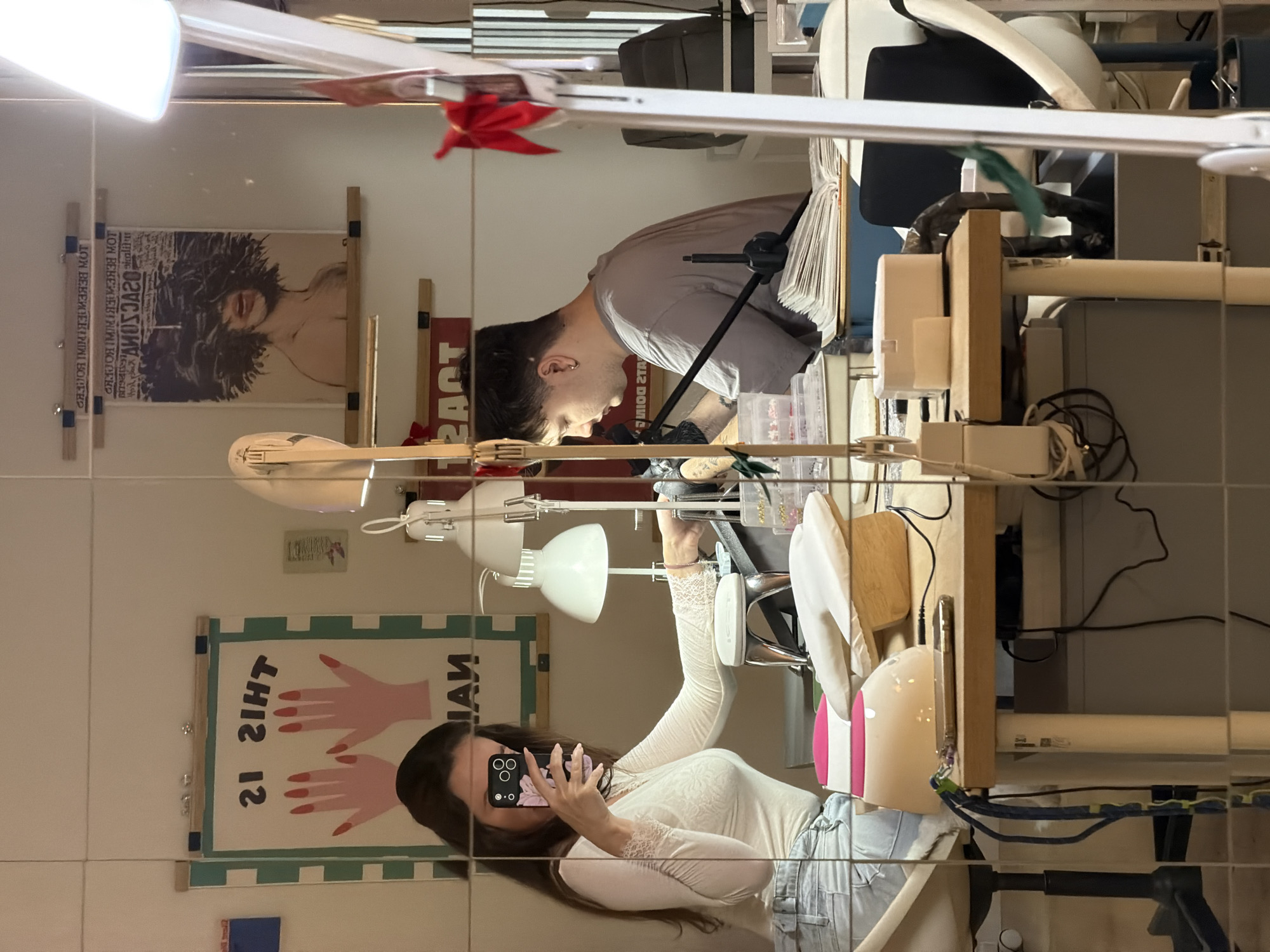 A mirrored image captures a woman with long brown hair, wearing a white lace-sleeved top and light-wash jeans, holding a phone. Reflected behind her is a man with short dark hair and a gray shirt working. In the background are framed posters. Overhead, a bright white lamp fixture illuminates the scene. The location is likely indoors, and while specific to Belgrade, Serbia is not directly discernible from the image.