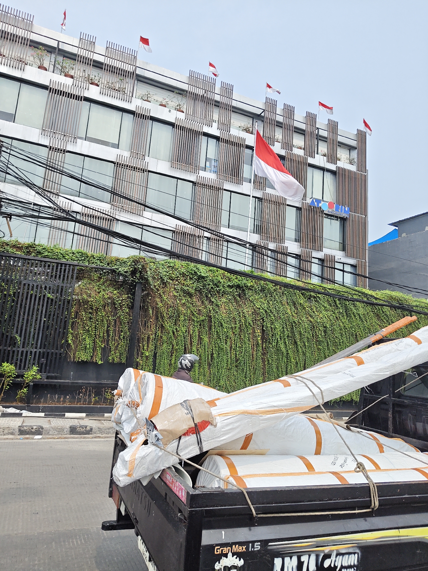 In West Jakarta, Indonesia, a flatbed truck is parked on a street, carrying a load of large, white, cylindrical objects wrapped in plastic and secured with orange tape and ropes. A person wearing a helmet is seated on the truck. Behind the truck, a modern building with glass windows and vertical wooden louvers is visible. Red and white Indonesian flags are displayed on the building. A dense green vine covers a black metal fence in front of the building. Electrical wires crisscross the scene in the foreground. The street surface is asphalt, with a sidewalk and a concrete barrier.