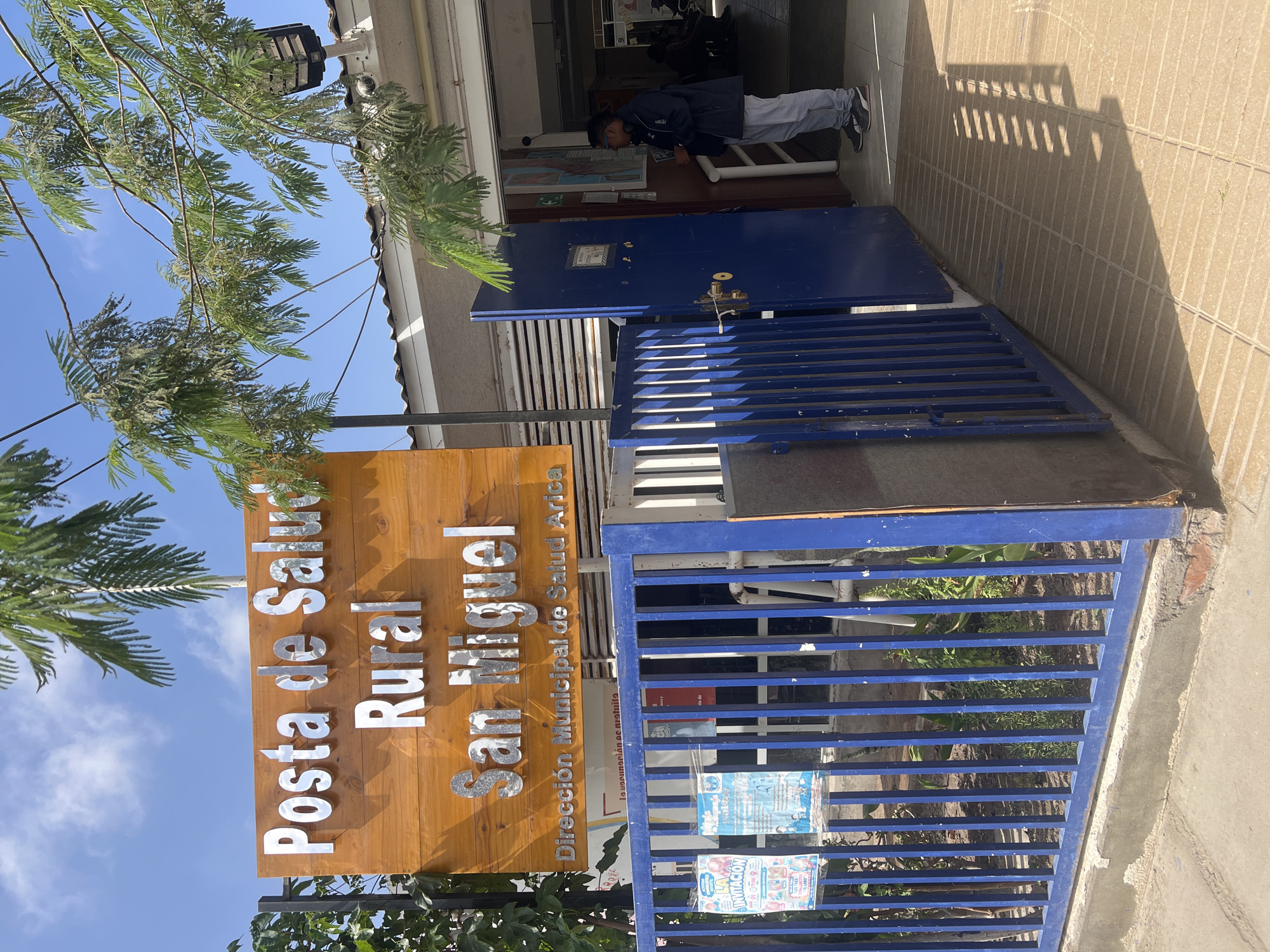 The image shows the entrance to a health post in a rural area called "San Miguel," located in Arica, Chile. The sign in front of the building reads "Posta de Salud Rural San Miguel" and "Dirección Municipal de Salud Arica," with "La vacunación es gratuita" written below. There is a blue metal gate and door partially closed in front of the entrance. A person is seen entering the building. The image is taken outdoors during the day, with trees visible in the foreground and blue sky above. There are informational flyers posted on the gate.