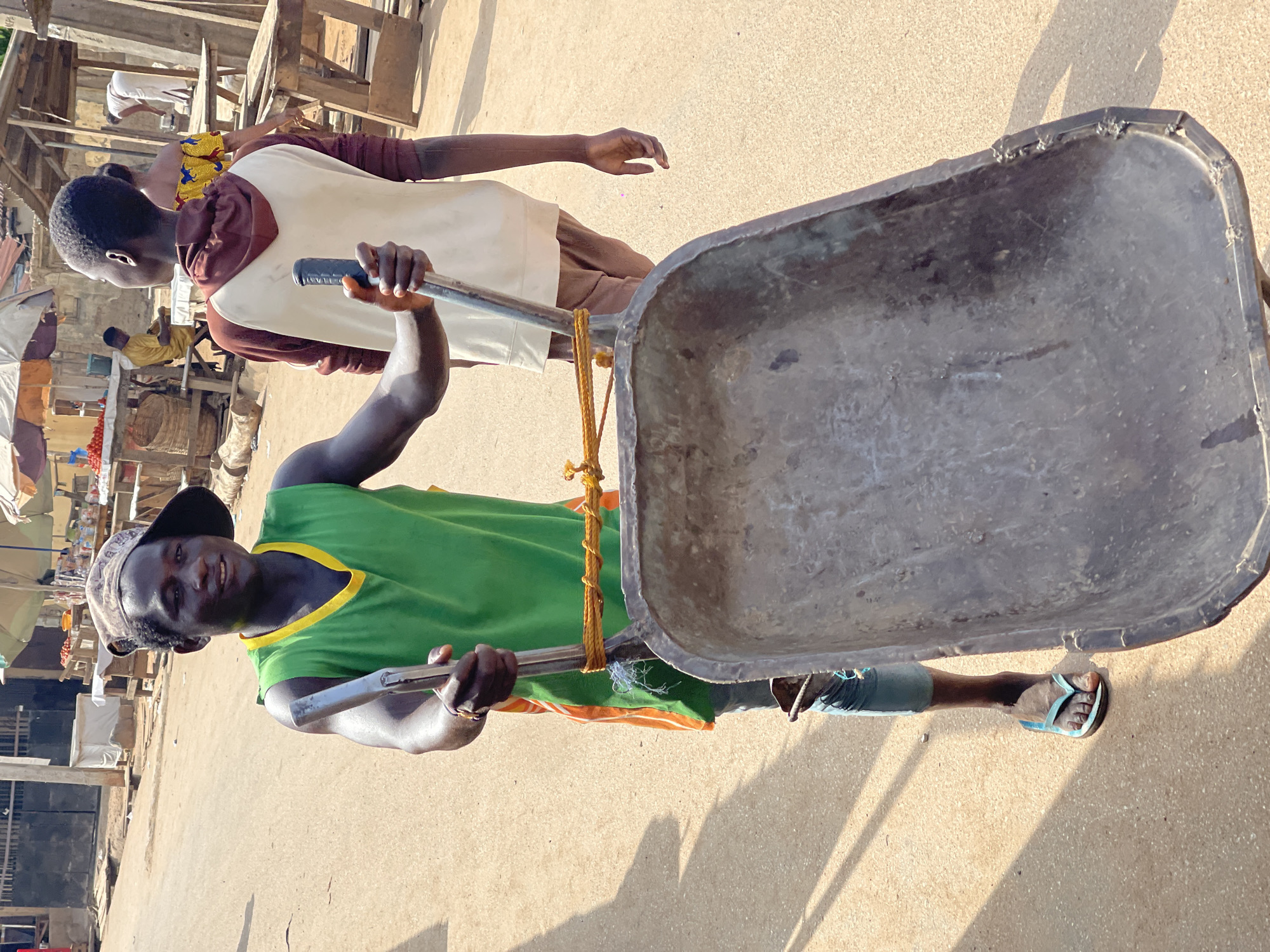 An outdoor scene in Kuchingoro, Nigeria, captures a bustling marketplace or street activity under strong sunlight, casting distinct shadows on the light-colored, dusty ground.
The primary subject is a dark-skinned man positioned centrally, looking towards the viewer with a slight smile. He wears a vibrant green sleeveless shirt with yellow trim, a patterned cap, and light blue flip-flops. He is actively holding the metal handles of a large, empty, well-used metal wheelbarrow, suggesting he is either pushing it or preparing to do so. A yellow rope is tied to one of the wheelbarrow handles.
Behind him and to the left stands another dark-skinned individual, appearing younger, dressed in a brown and white long-sleeved top and brown trousers. This person looks to the left, away from the viewer.
In the background, the setting is clearly a market or an area with street vendors. Various wooden structures, stalls, and tables are visible, some appearing to display goods. One stall holds baskets or containers, possibly filled with produce like tomatoes. Other indistinct figures can be seen further back amidst the market infrastructure, including an umbrella or canopy, providing shade. No visible text is present in the image.
