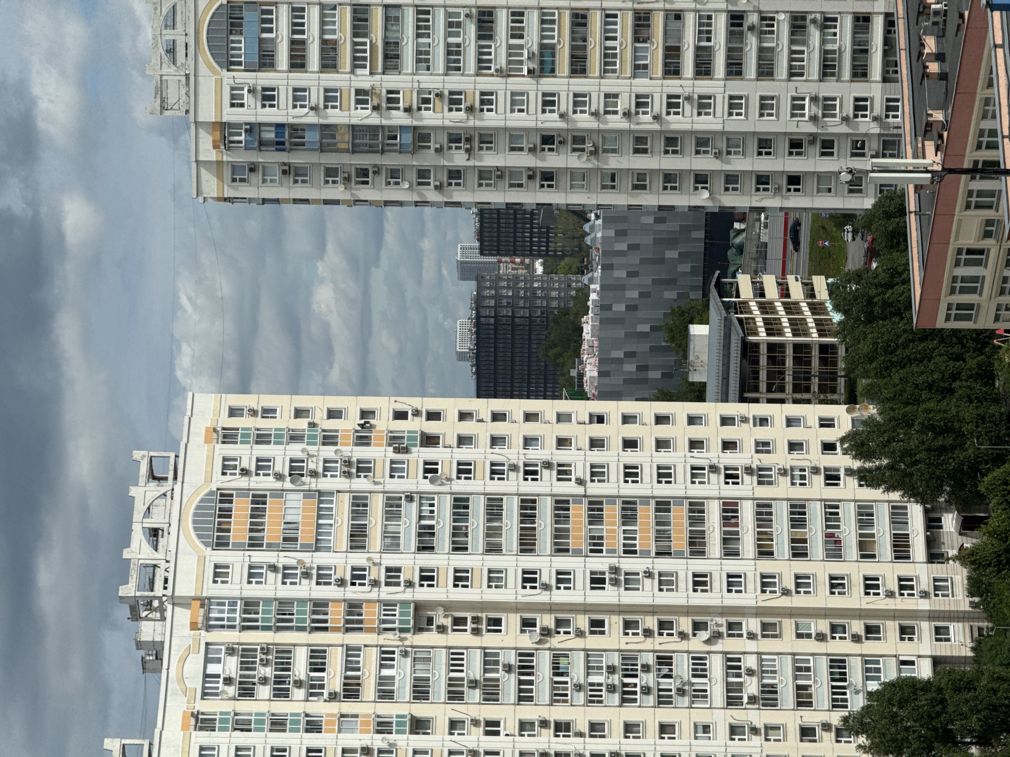 The image depicts a view of a residential area in Moscow, Russia, characterized by several high-rise apartment buildings. The foreground features two prominent apartment buildings, one on the left and one on the right, both appearing to be of similar architectural style with numerous windows and balconies. The building on the left is lighter in color, possibly beige or cream, with accent colors of green and orange around some windows. The building on the right has a similar color scheme, with a mix of beige, white, and blue accents. Both buildings have many visible air conditioning units mounted on their facades.
In the middle ground, several other buildings are visible, including darker, more modern-looking high-rise structures that contrast with the older apartment buildings. There is also a building with a gray, textured facade and another multi-story building with visible internal structures. Some trees are scattered throughout the scene, particularly in front of the middle-ground buildings.
The background shows a cloudy sky with dark, heavy clouds, suggesting overcast weather. In the distance, more buildings can be seen, indicative of a dense urban environment. A road with at least one car is also visible in the middle ground, suggesting active transportation routes within the neighborhood. The overall scene conveys a sense of a densely populated urban landscape with a mix of older and newer residential architecture.